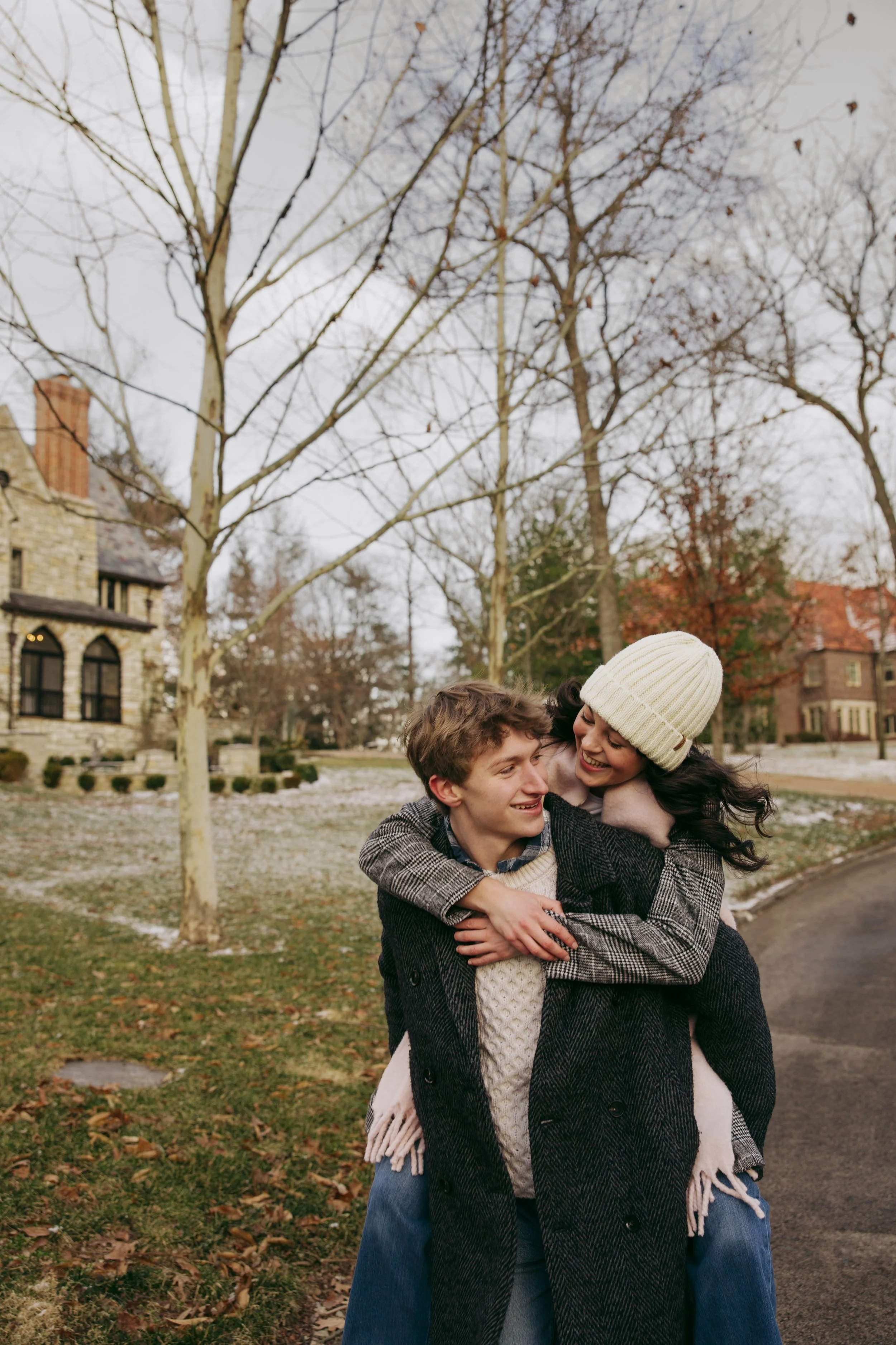 A happy couple embracing outdoors on a fall day, with trees and historic houses in the background, autumn leaves scattered on the ground.