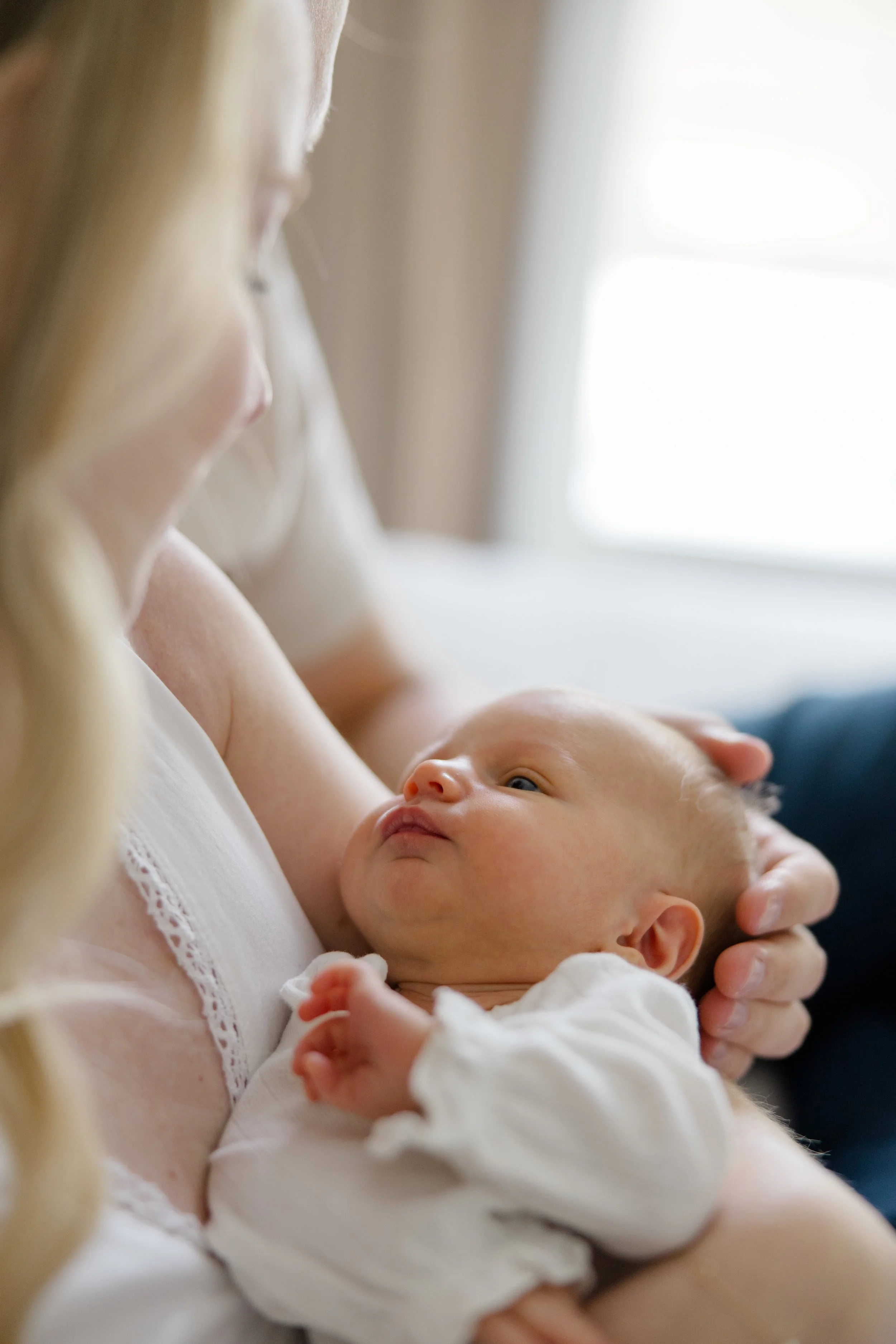 A woman holding a newborn baby close to her chest, the baby looking up at her with wide eyes. The woman wears a white top, and the setting appears to be indoors near a window with bright light.
