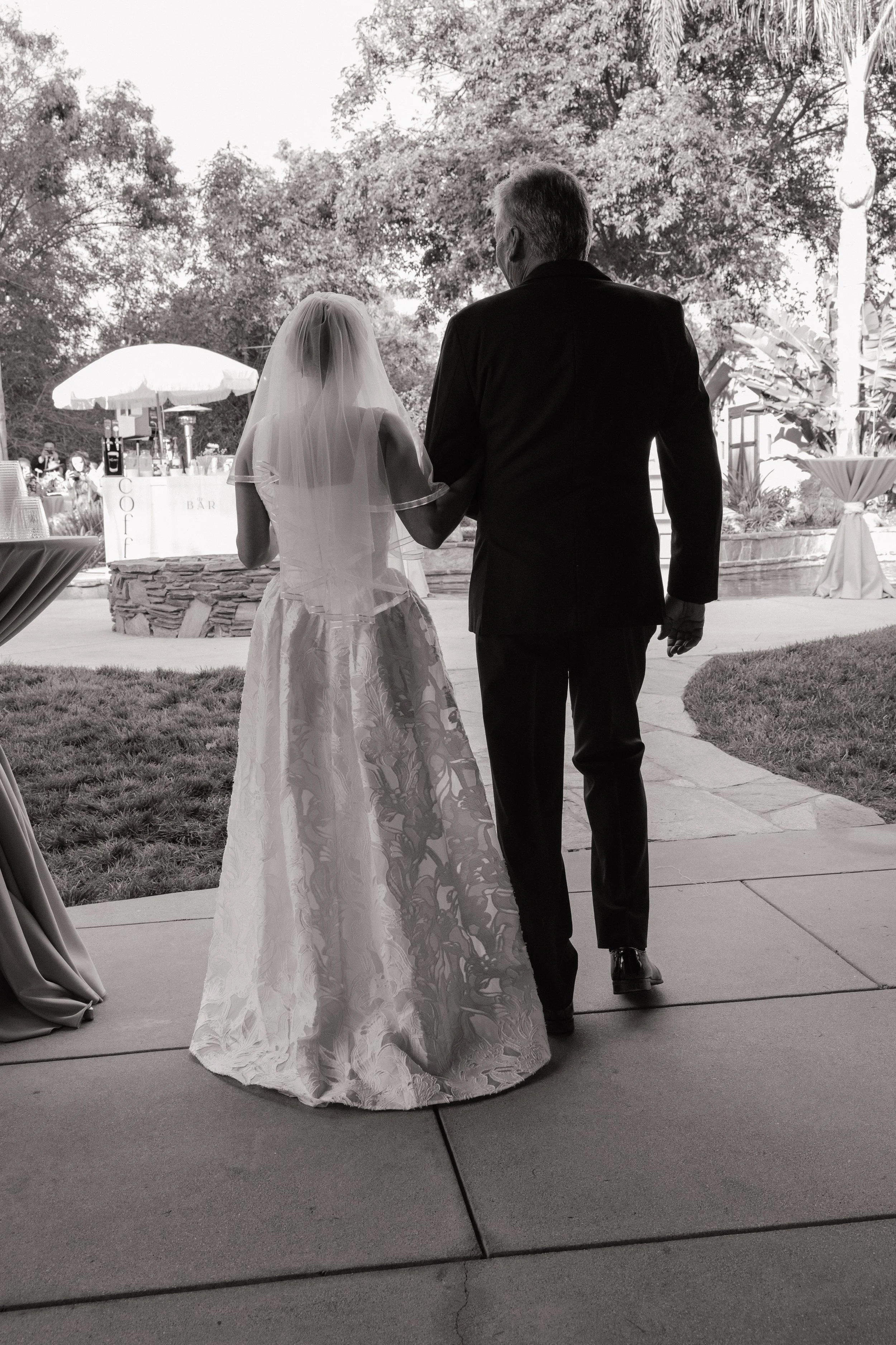 A bride and an older man walking outside at a wedding reception. The bride is wearing a lace wedding gown and veil, holding the older man's hand. The scene is set outdoors near tables and an umbrella, with trees in the background.