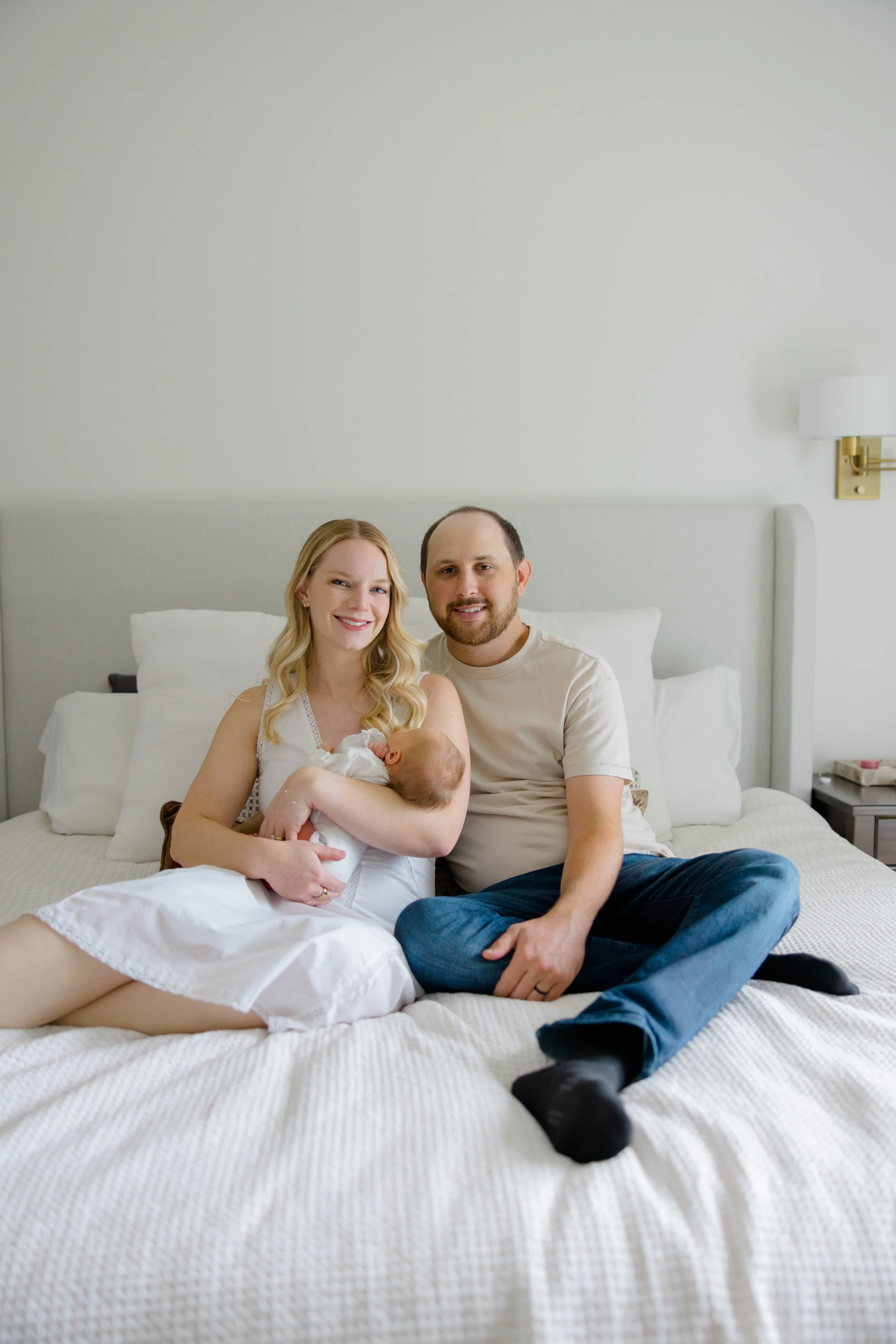 A smiling woman with blonde hair holding a newborn baby, sitting on a bed with a man, in a bright bedroom with white walls and modern decor.