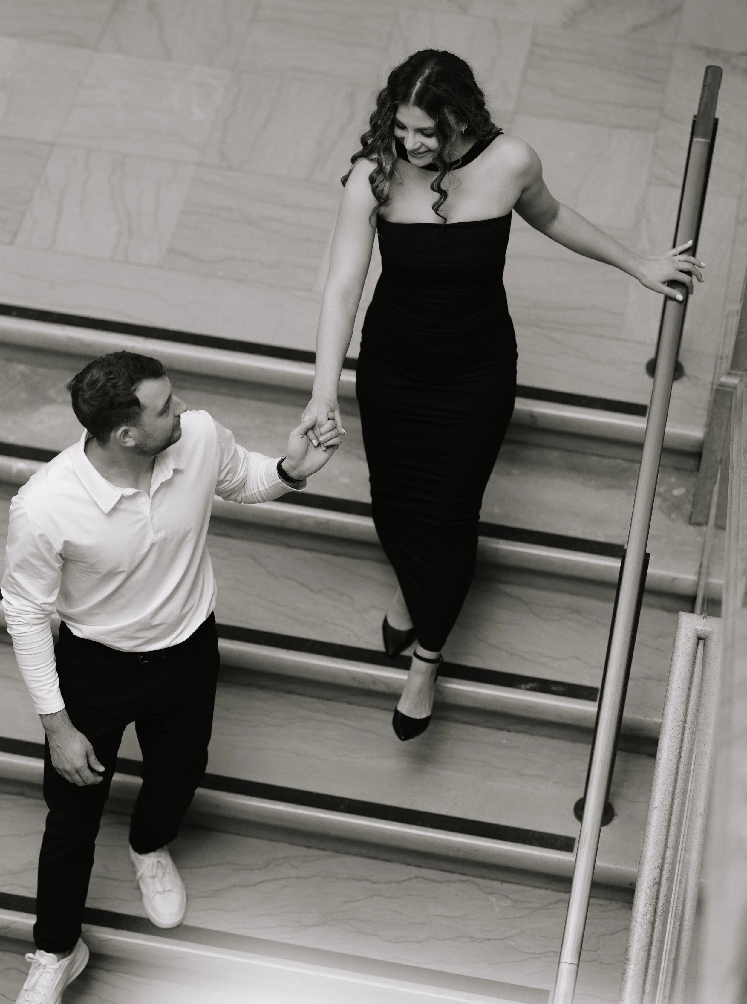 A woman in a black dress and high heels leaning against a railing, holding hands with a man kneeling on one knee, inside a building with marble walls and stairs.