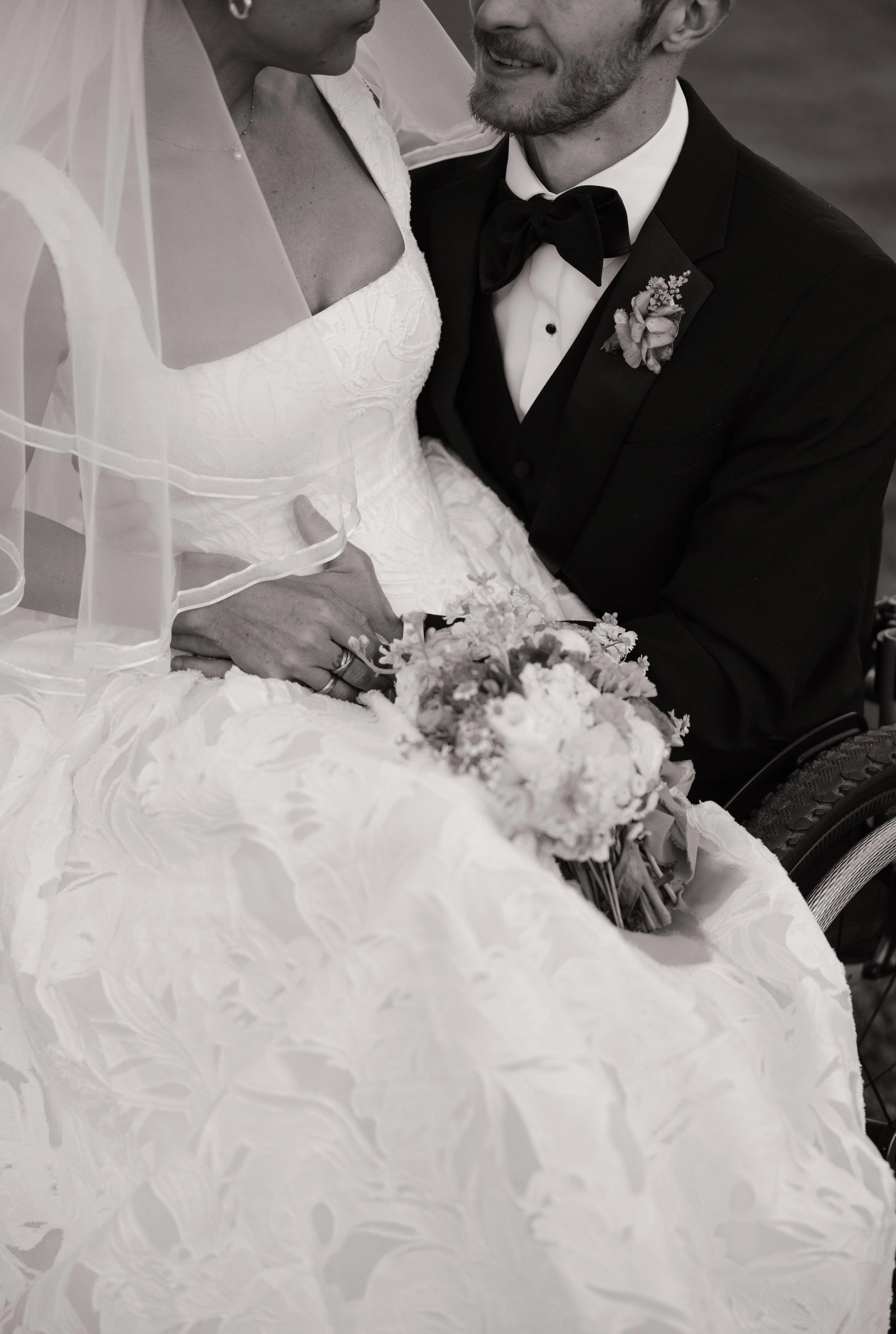 A black-and-white photograph of a couple on their wedding day, with the bride in a white lace wedding dress and veil, holding a bouquet of flowers, and the groom in a suit with a bow tie and a boutonniere, close together, smiling at each other.