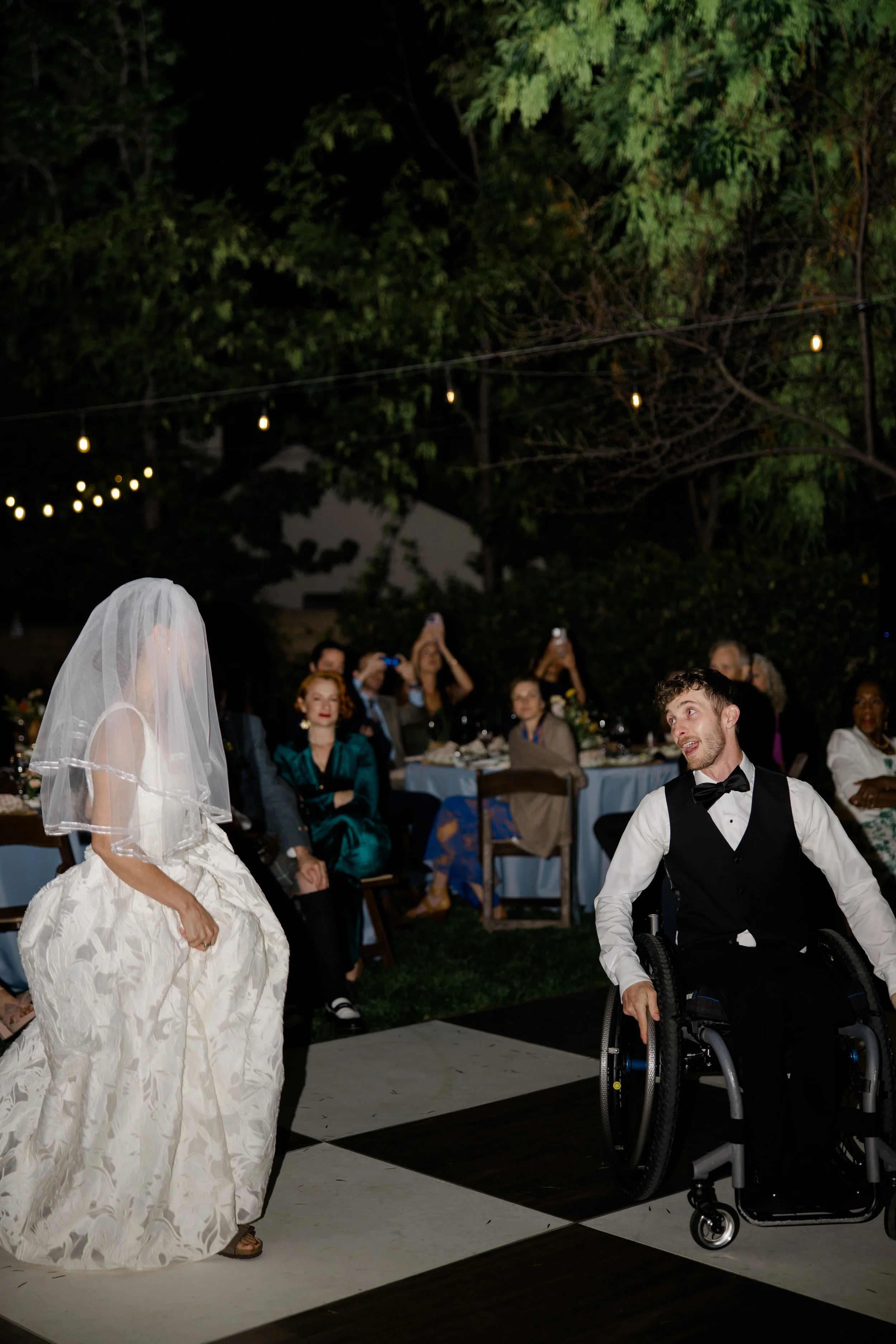 A bride and groom dance at their outdoor wedding reception at night, with guests sitting at decorated tables and string lights overhead.