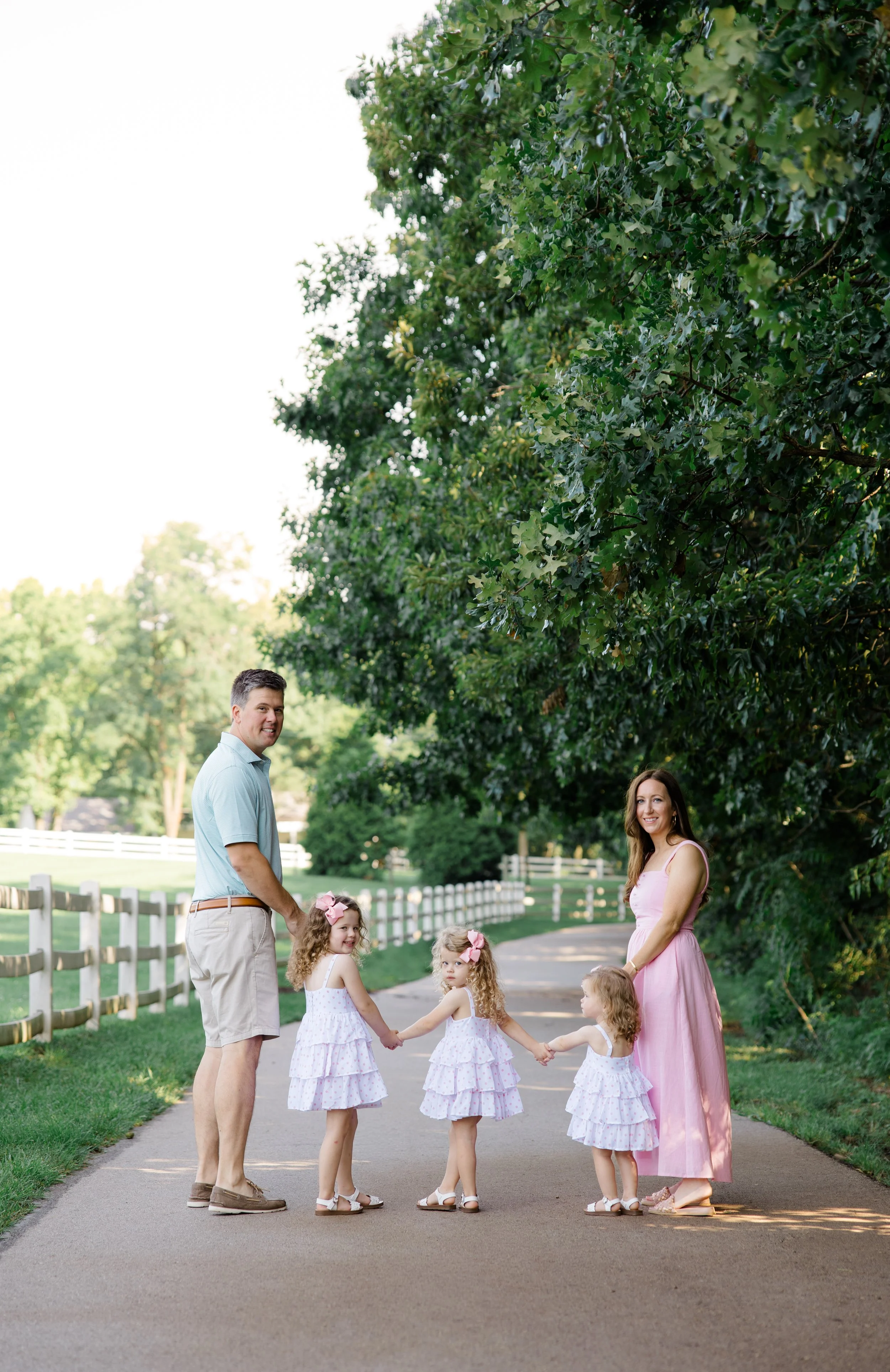 A family of four with three young girls holding hands and smiling on a park pathway surrounded by lush green trees and a white fence.