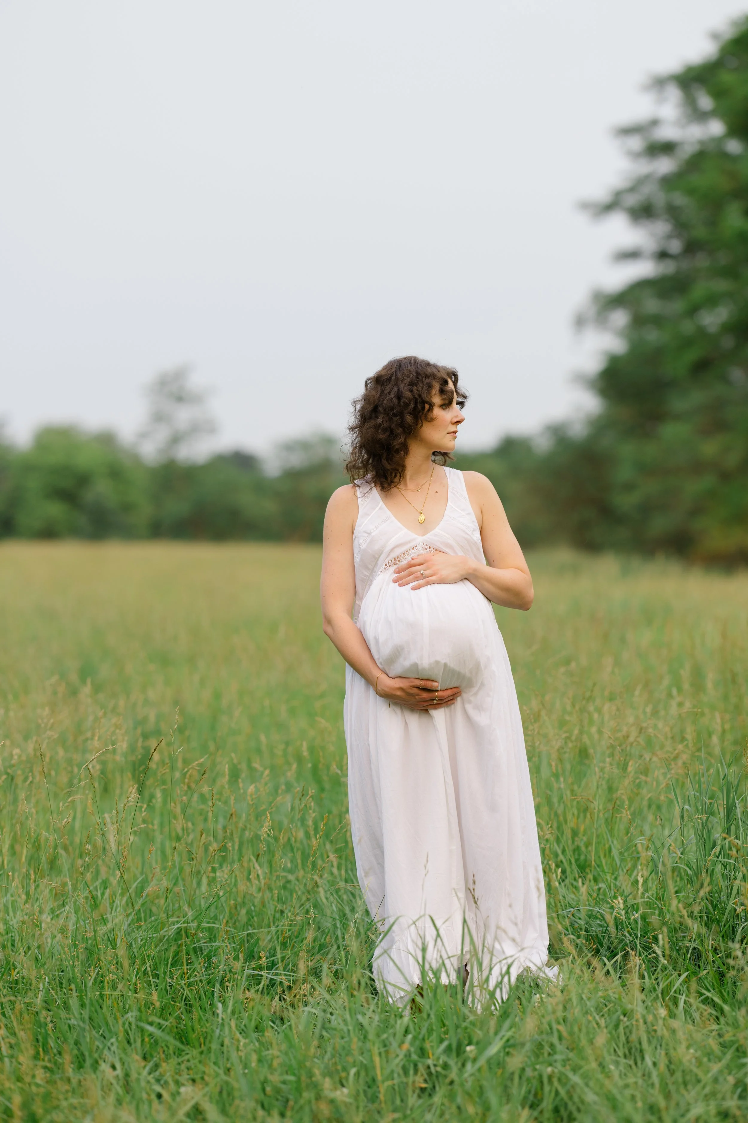 A pregnant woman with curly brown hair stands in a green field, wearing a white dress and holding her belly, with trees in the background.