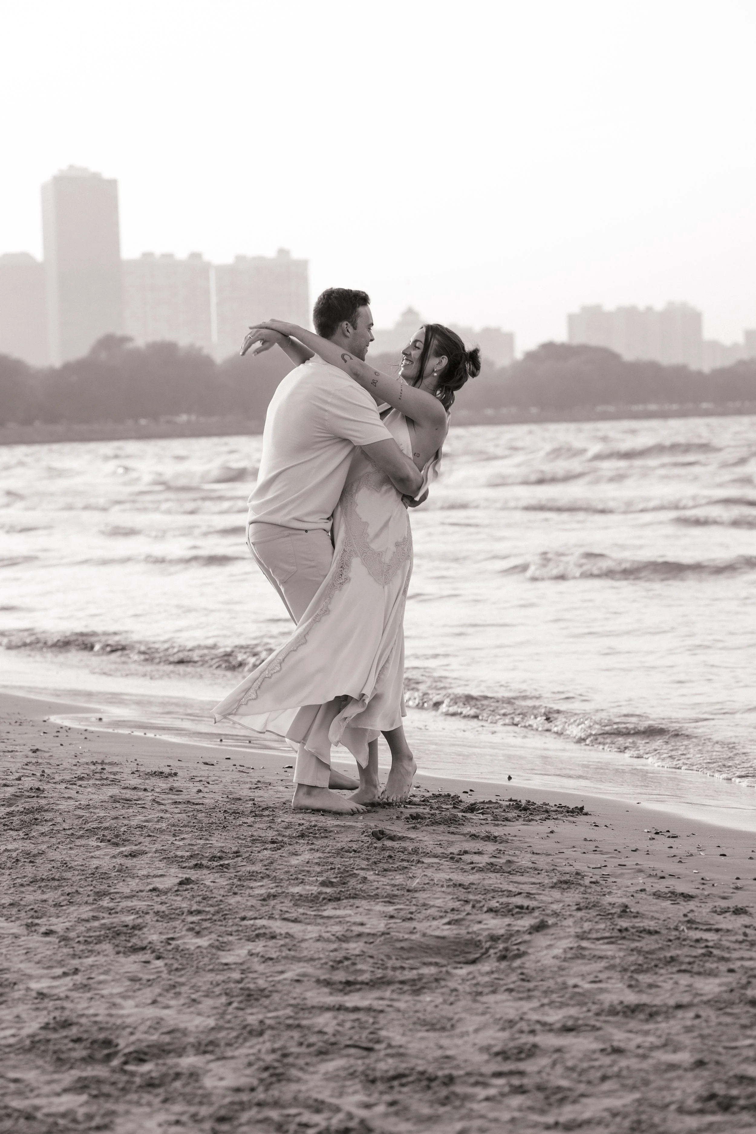 A couple dancing barefoot on the beach with the city skyline in the background, captured in black and white.