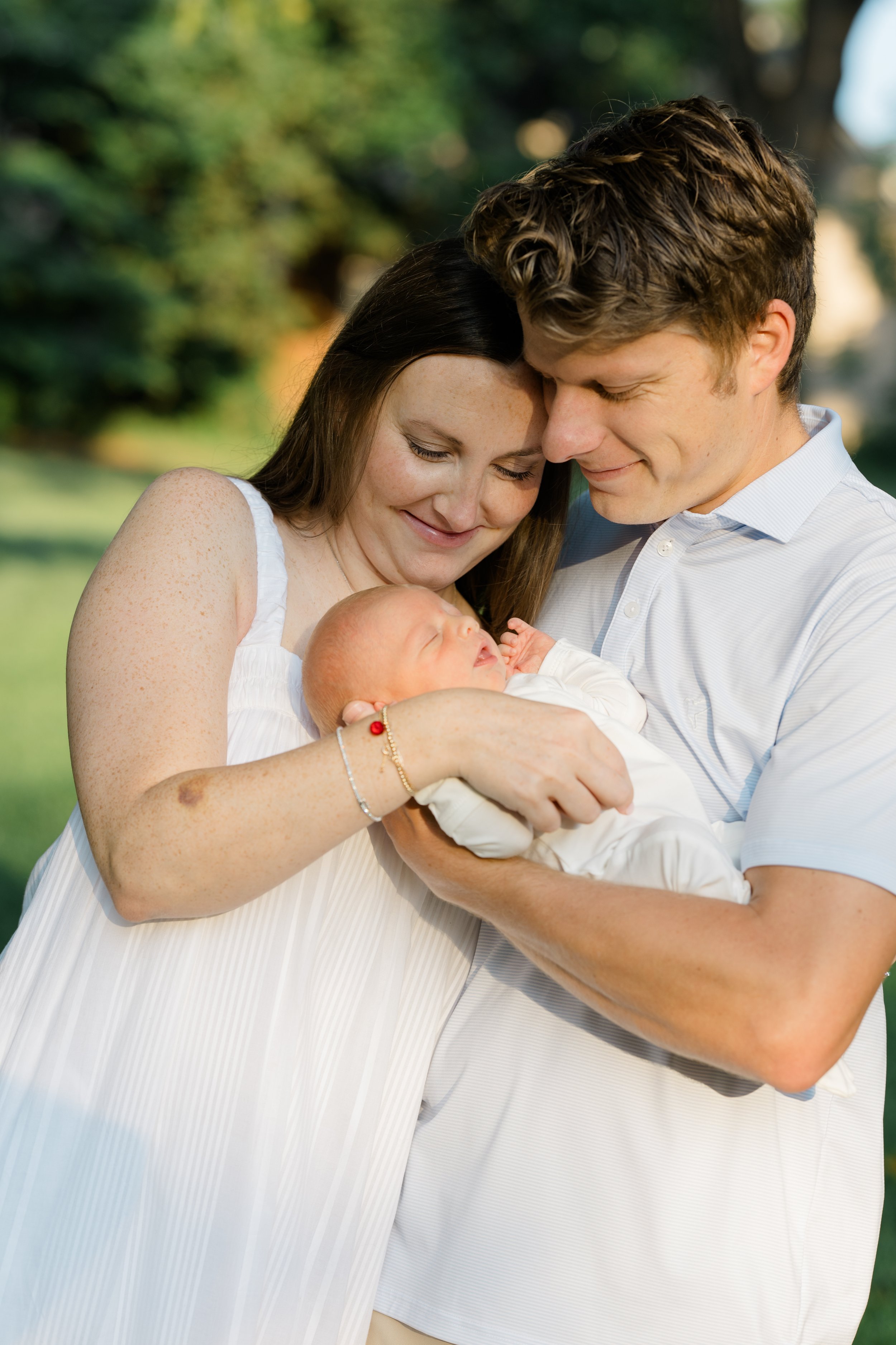 A happy couple holding a newborn baby outdoors, smiling and touching foreheads, with a lush green background.