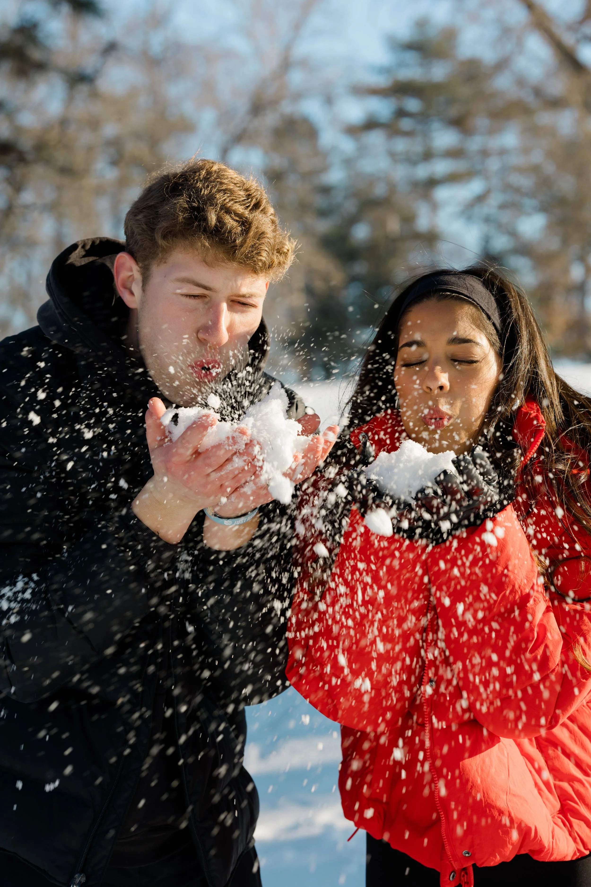 A young man and woman playing in the snow, blowing snow from their hands with their eyes closed, outdoors in a winter landscape with trees.