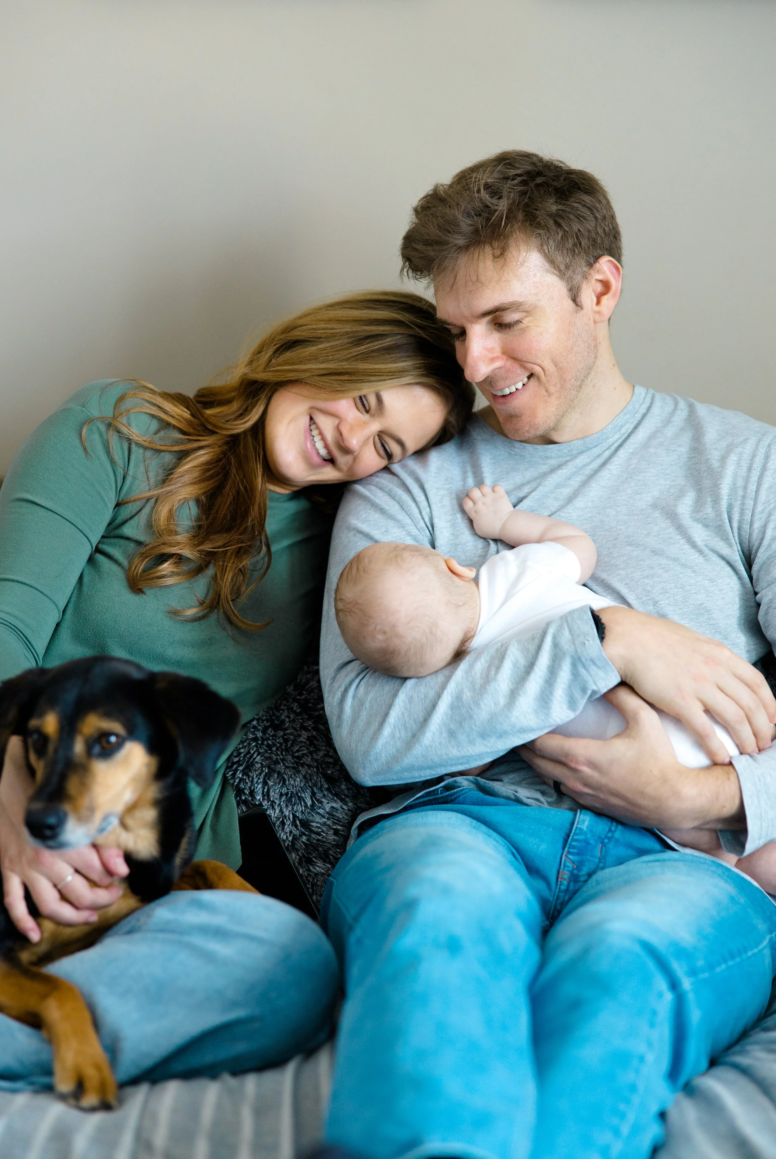 A happy family with a man, woman, baby, and dog sitting together on a couch. The woman and man are smiling, the woman resting her head on the man's shoulder, and the baby is being held by the man. The dog is sitting in the foreground.