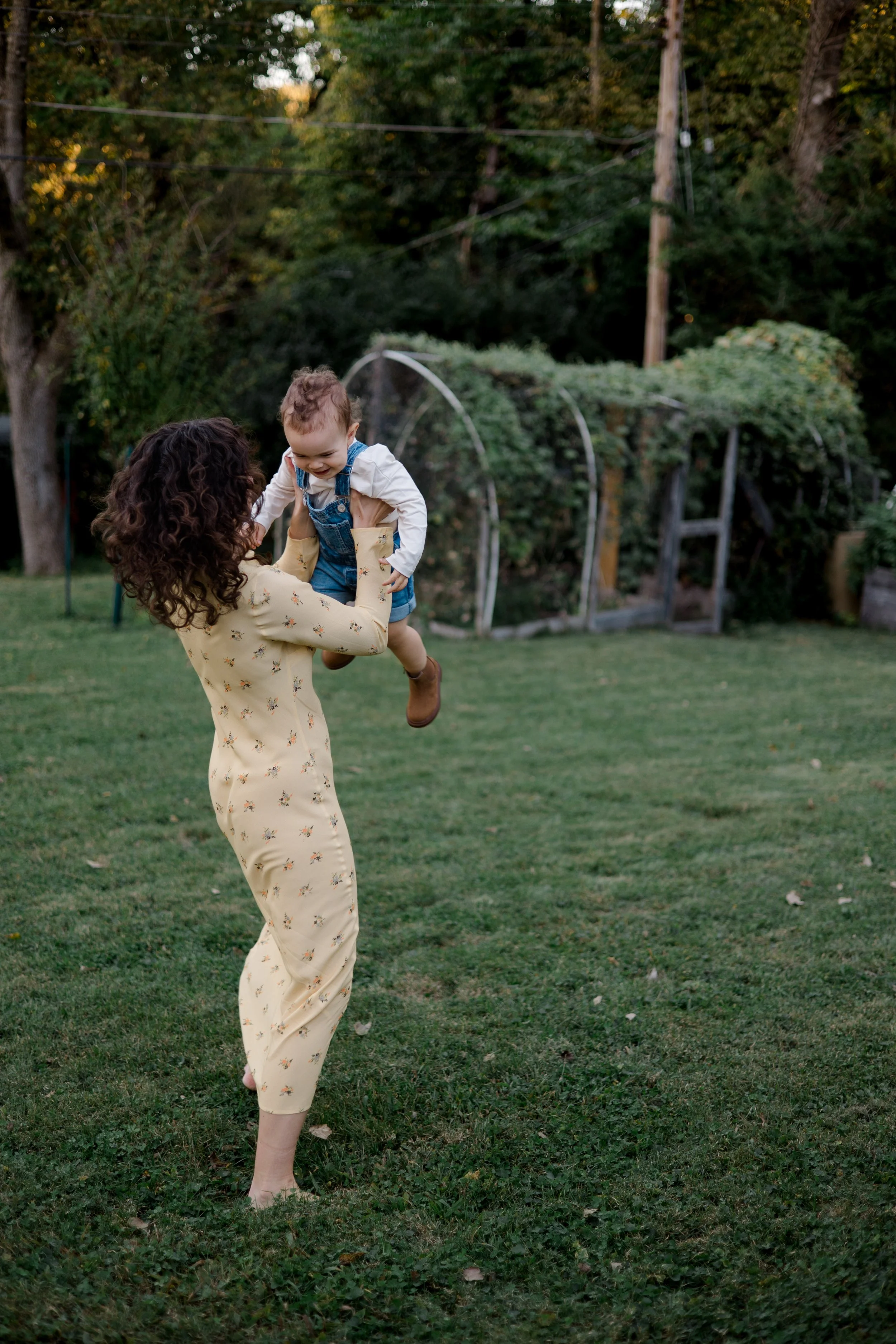 A woman with curly hair is playing with a young child in a backyard with green grass and trees. She is lifting the child, who is smiling and wearing overalls, into the air.