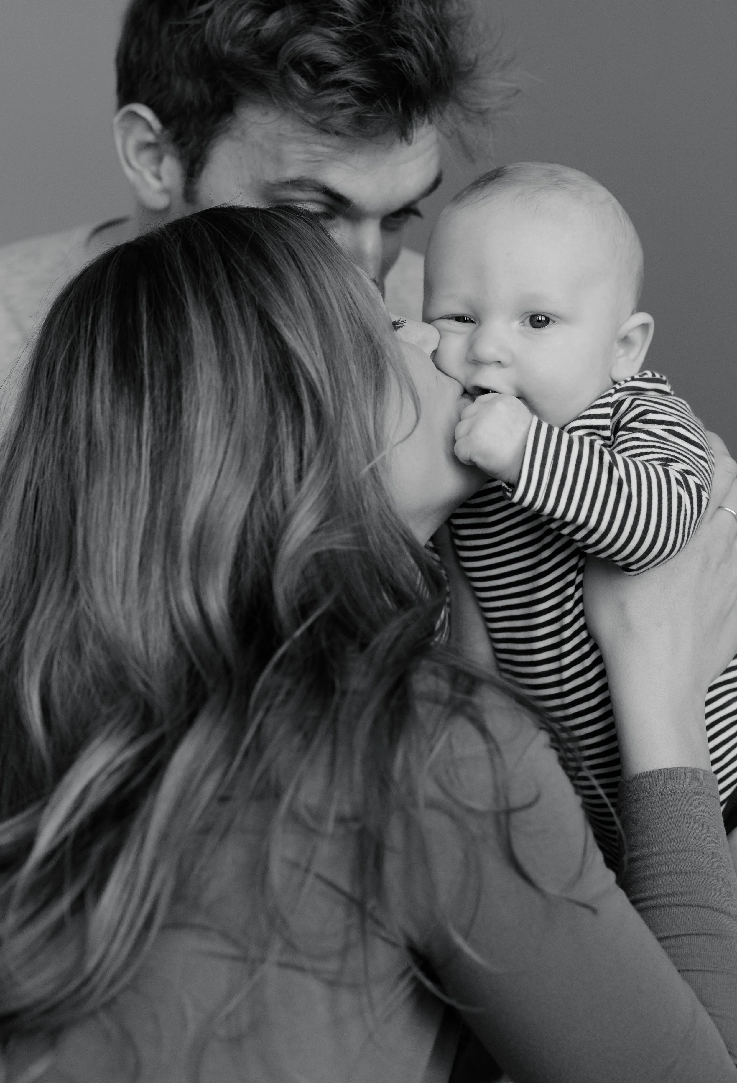 A woman with long, wavy hair kissing a baby with a striped shirt on the cheek, with a man leaning in close from above, all in black and white.