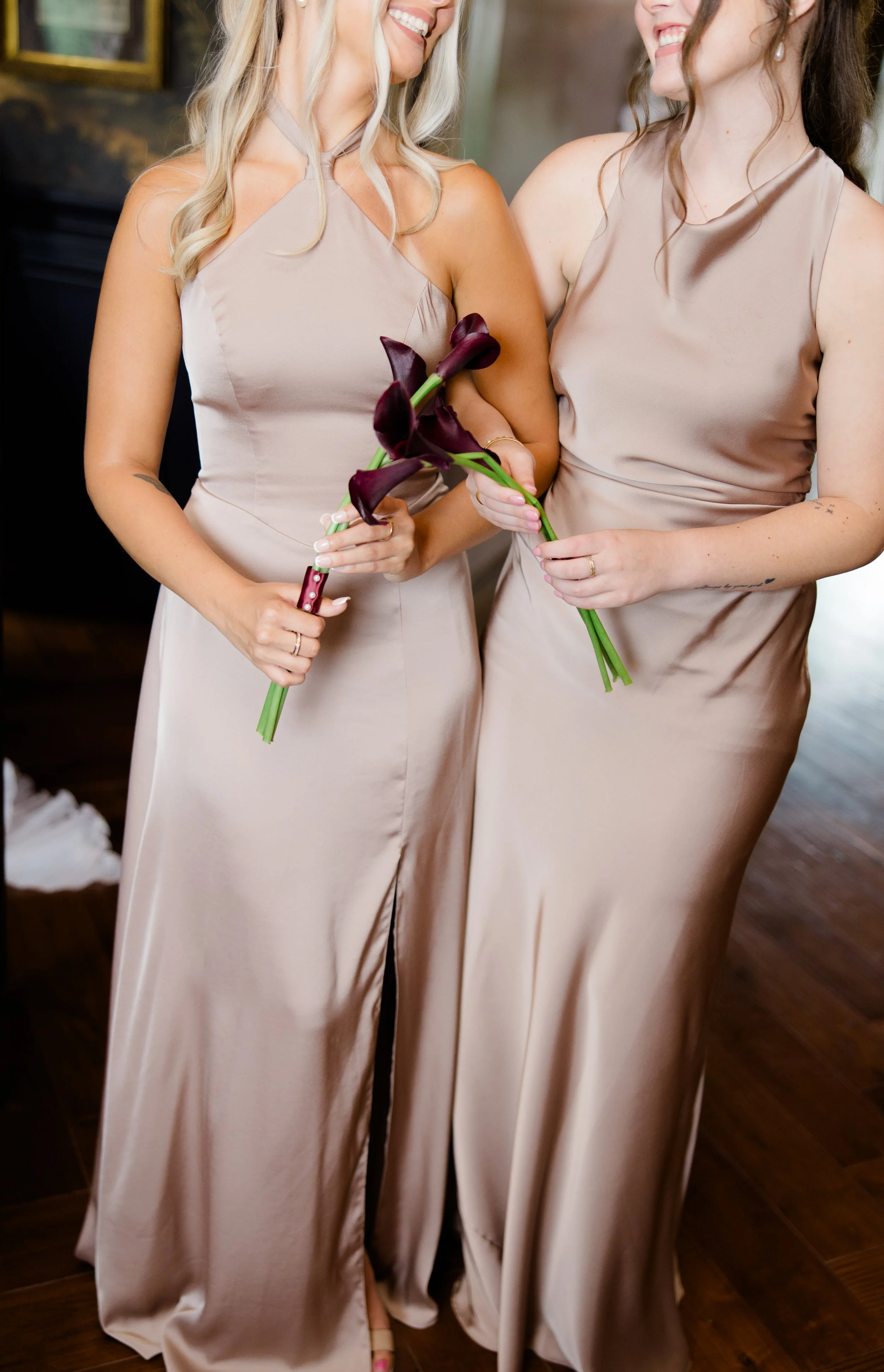 Two women wearing matching satin beige dresses holding dark purple calla lily flowers, smiling at each other, in an indoor setting.