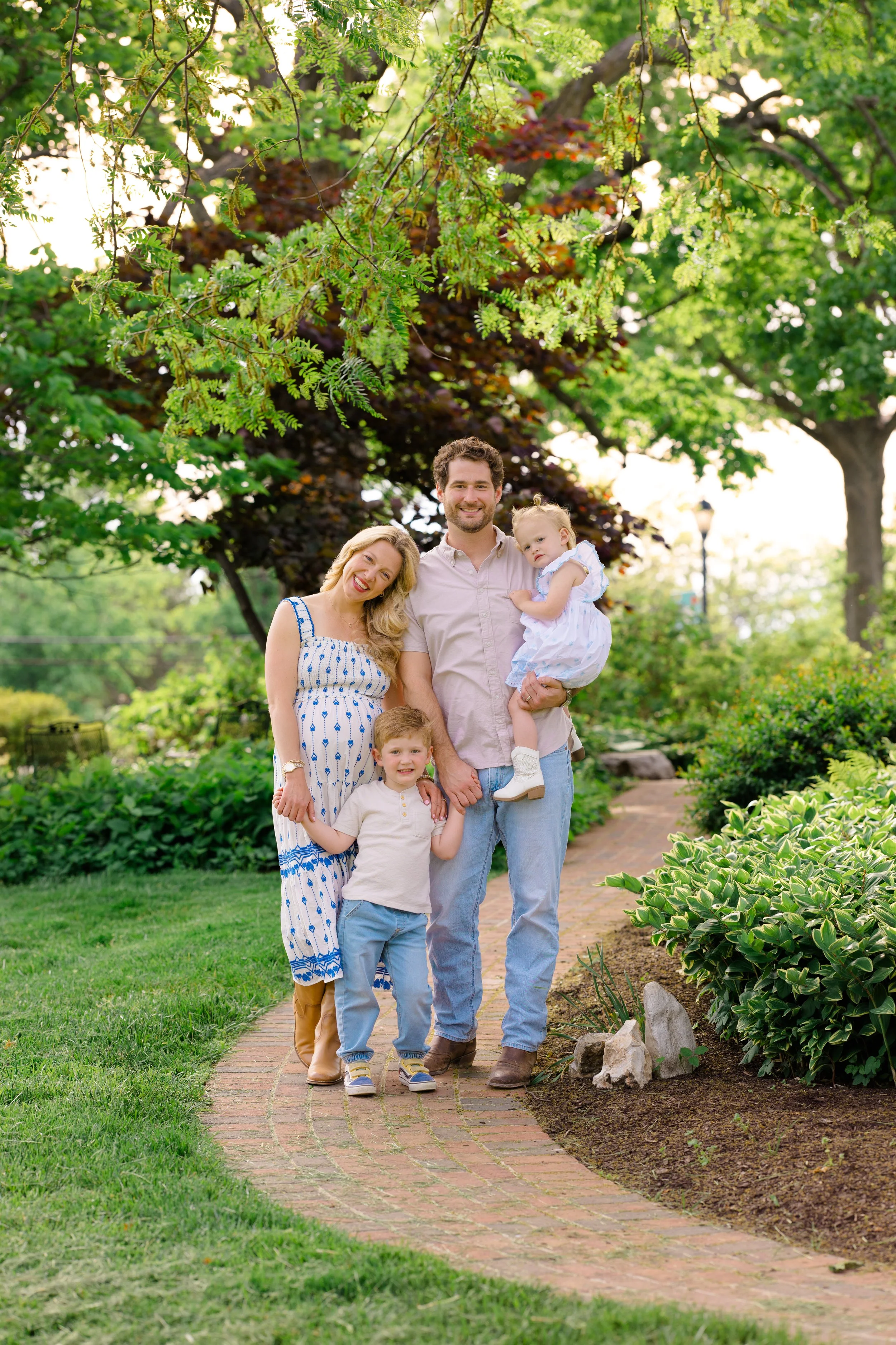 A family of five, including a man, a woman, two boys, and a girl, walking on a brick path in a lush green garden. The man is holding the girl, while the woman and the boys walk beside him, smiling and enjoying the outdoors. There are trees and bushes