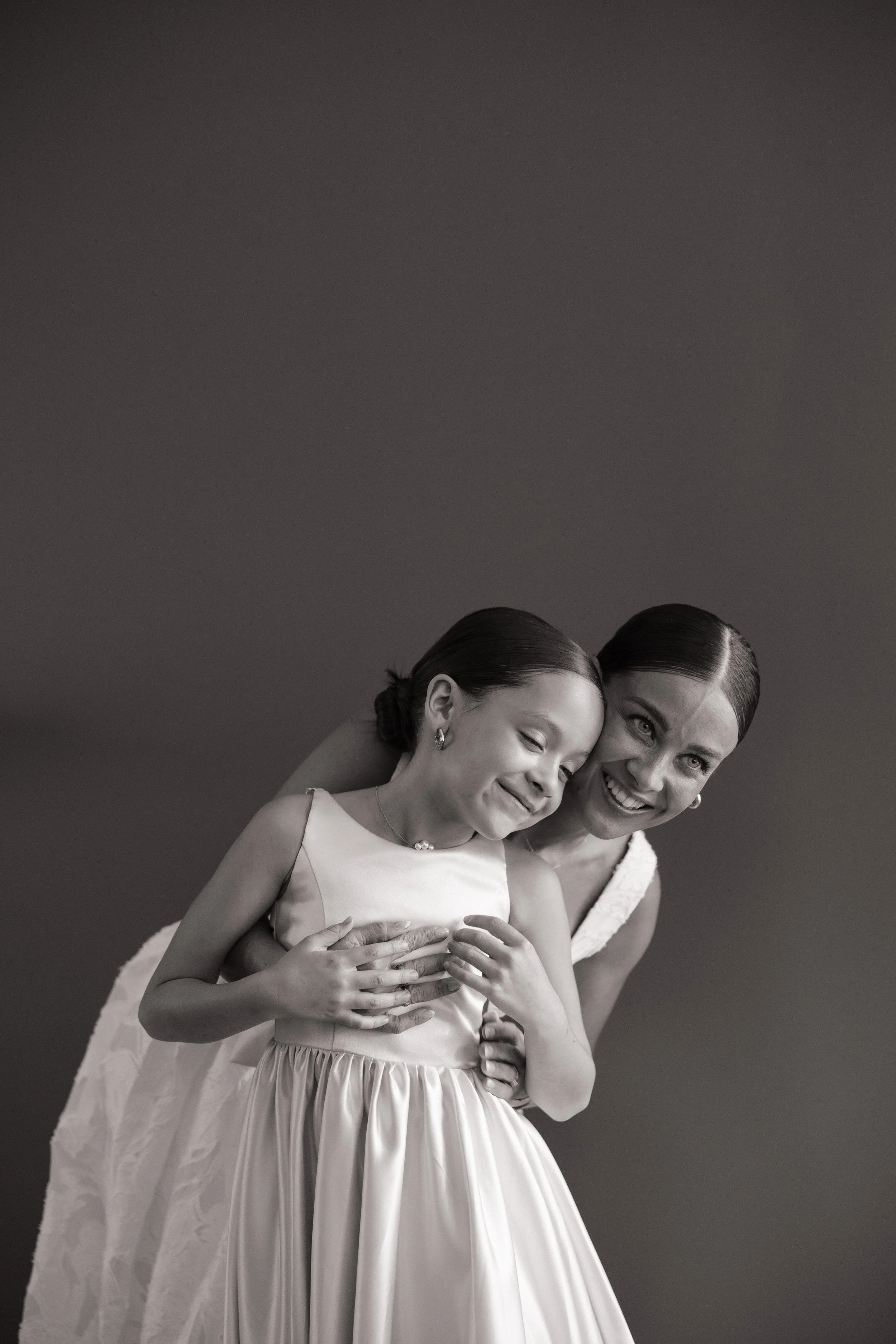 A black and white photo of a woman and girl in formal dresses hugging and smiling, looking at the camera, with dark plain background.