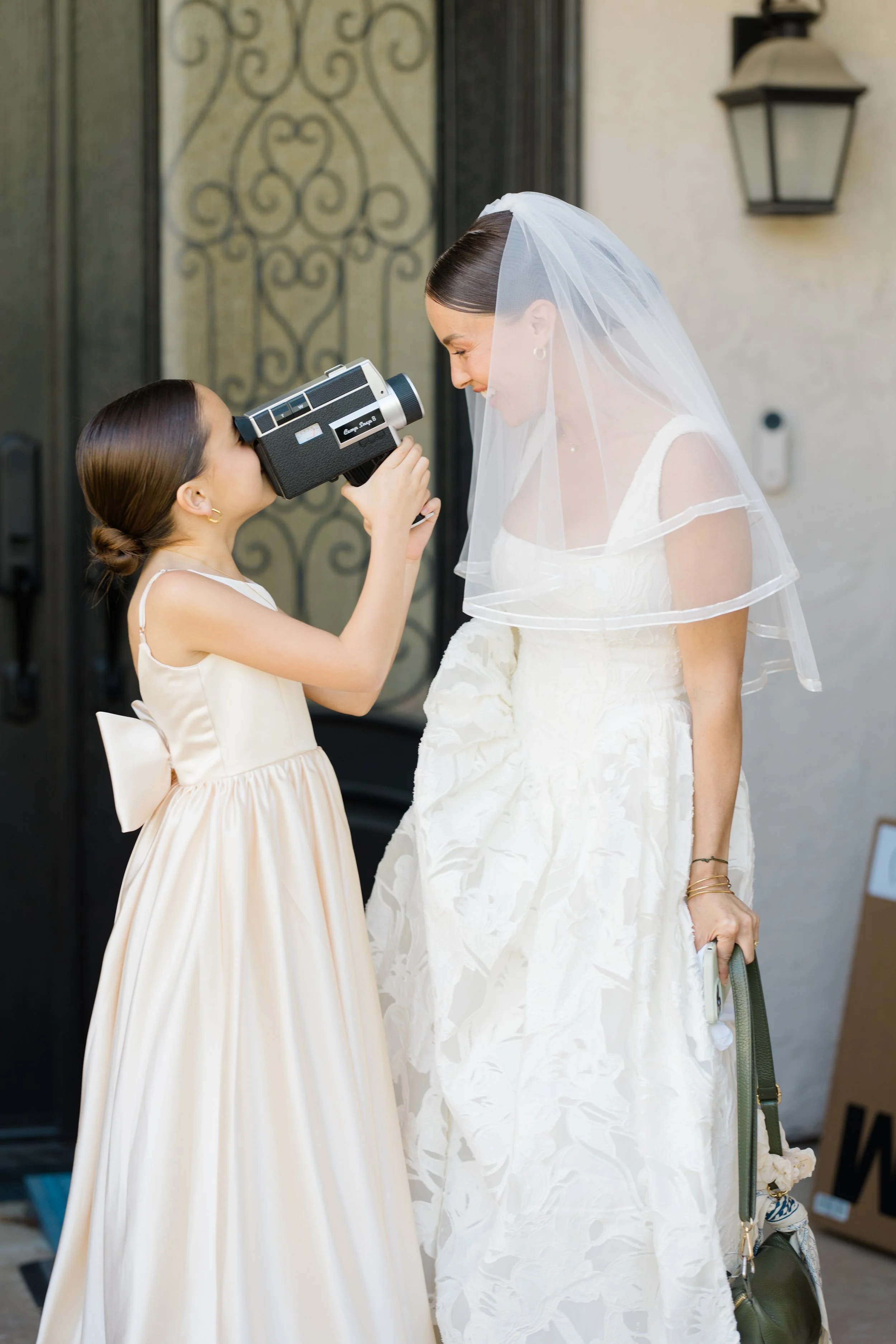 A young girl in a cream dress holding a scanner or camera to a bride's forehead. The bride is dressed in a white wedding gown with a tulle veil, and is smiling as the girl scans her forehead. They are standing outdoors near a decorative gate and a wa