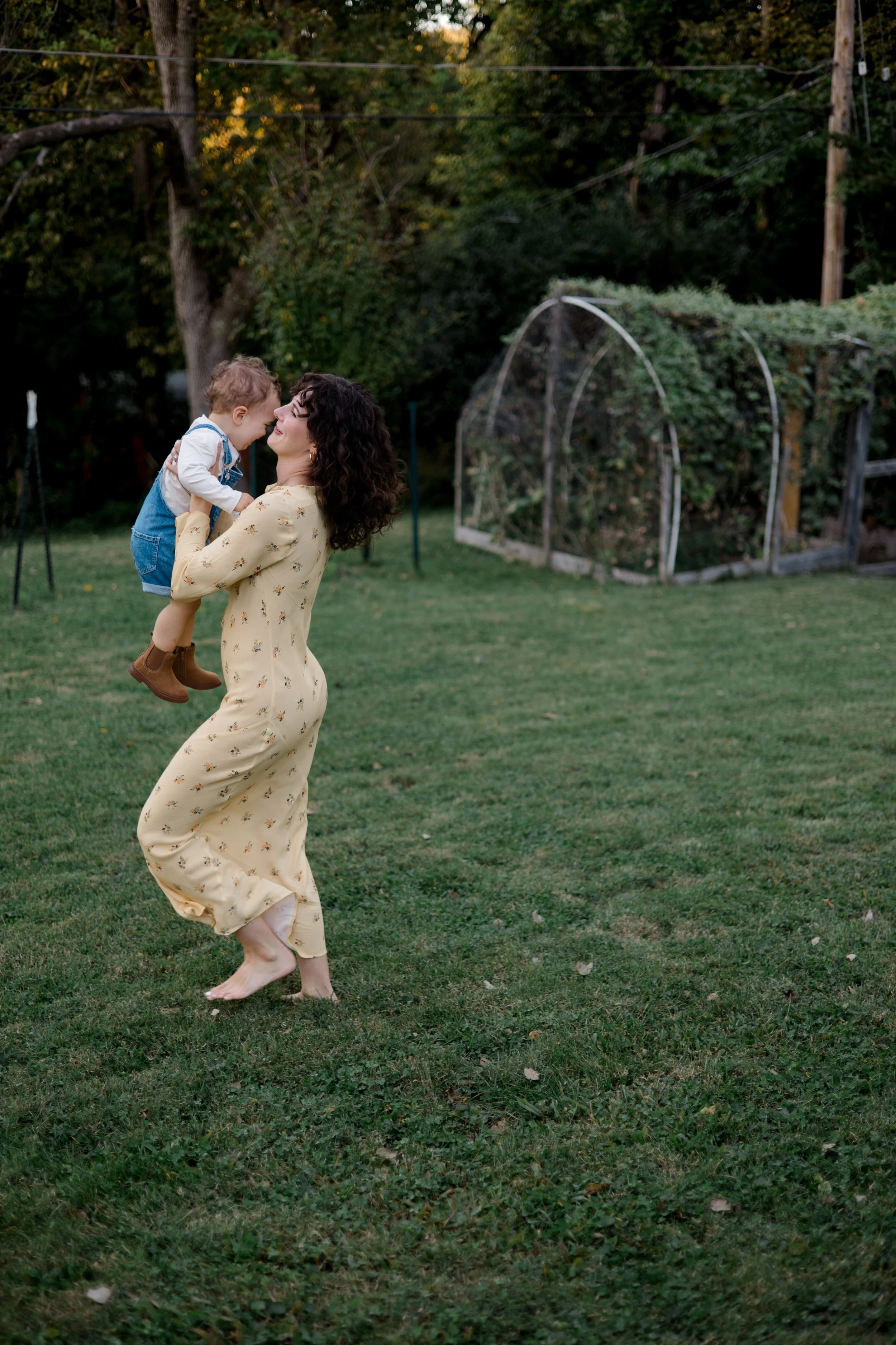 A woman in a yellow floral dress and barefoot is joyfully spinning outdoors while holding a young child wearing a white shirt and blue denim overalls.