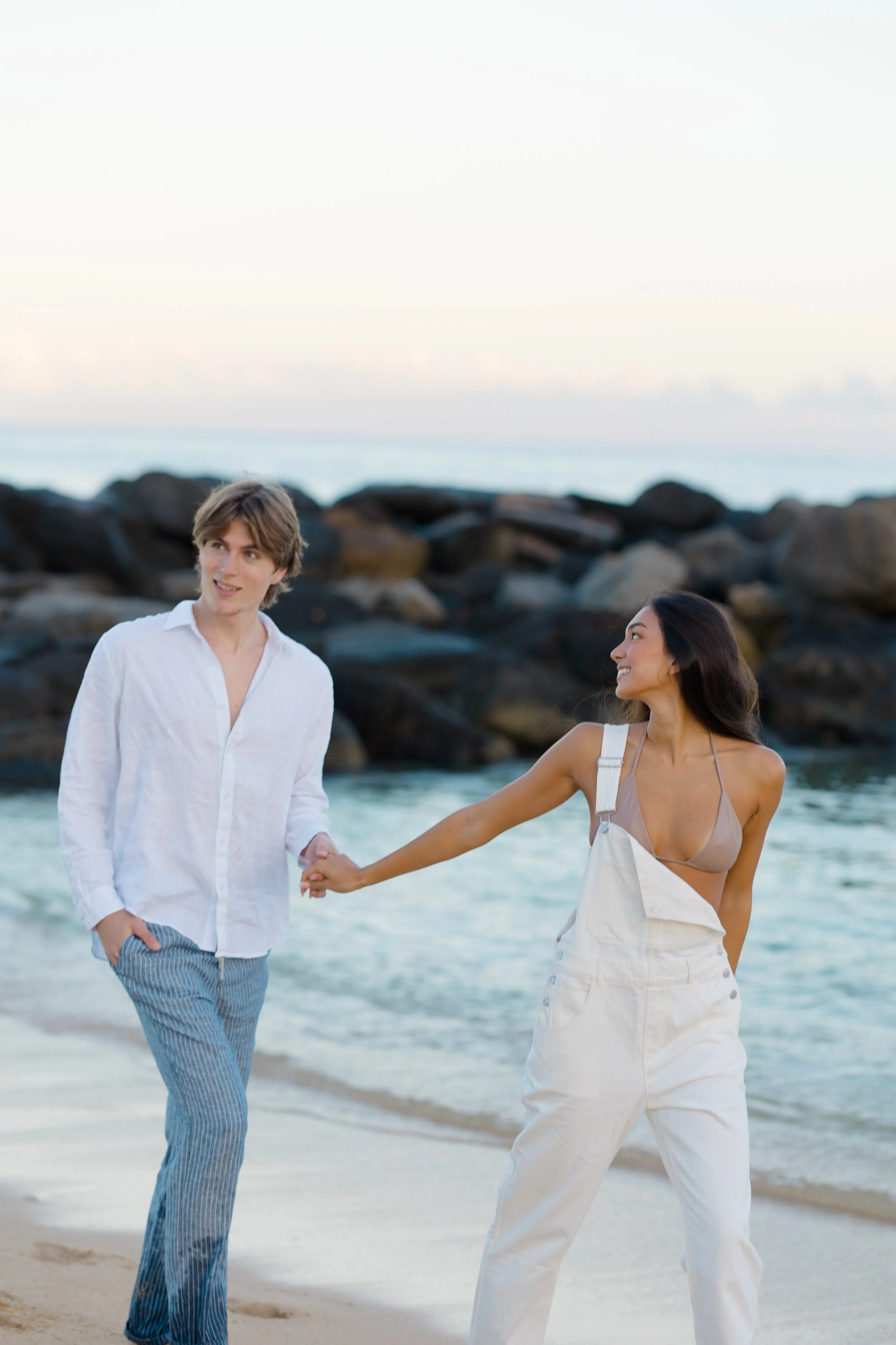 A young man and woman holding hands and walking on a beach with rocks in the background. The man is dressed in a white shirt and striped pants, and the woman is wearing a beige sleeveless top and light-colored overalls.