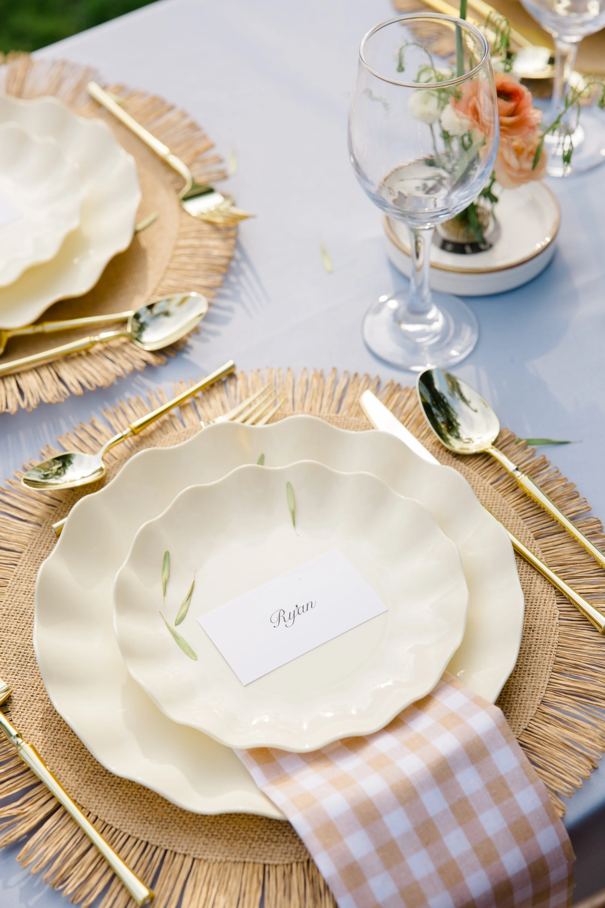 Elegant outdoor table setting with a white scalloped plate, a place card with the name "Ryan," gold utensils, and a wine glass, all on a round woven placemat with a gingham napkin nearby, decorated with pink and white flowers in the background.