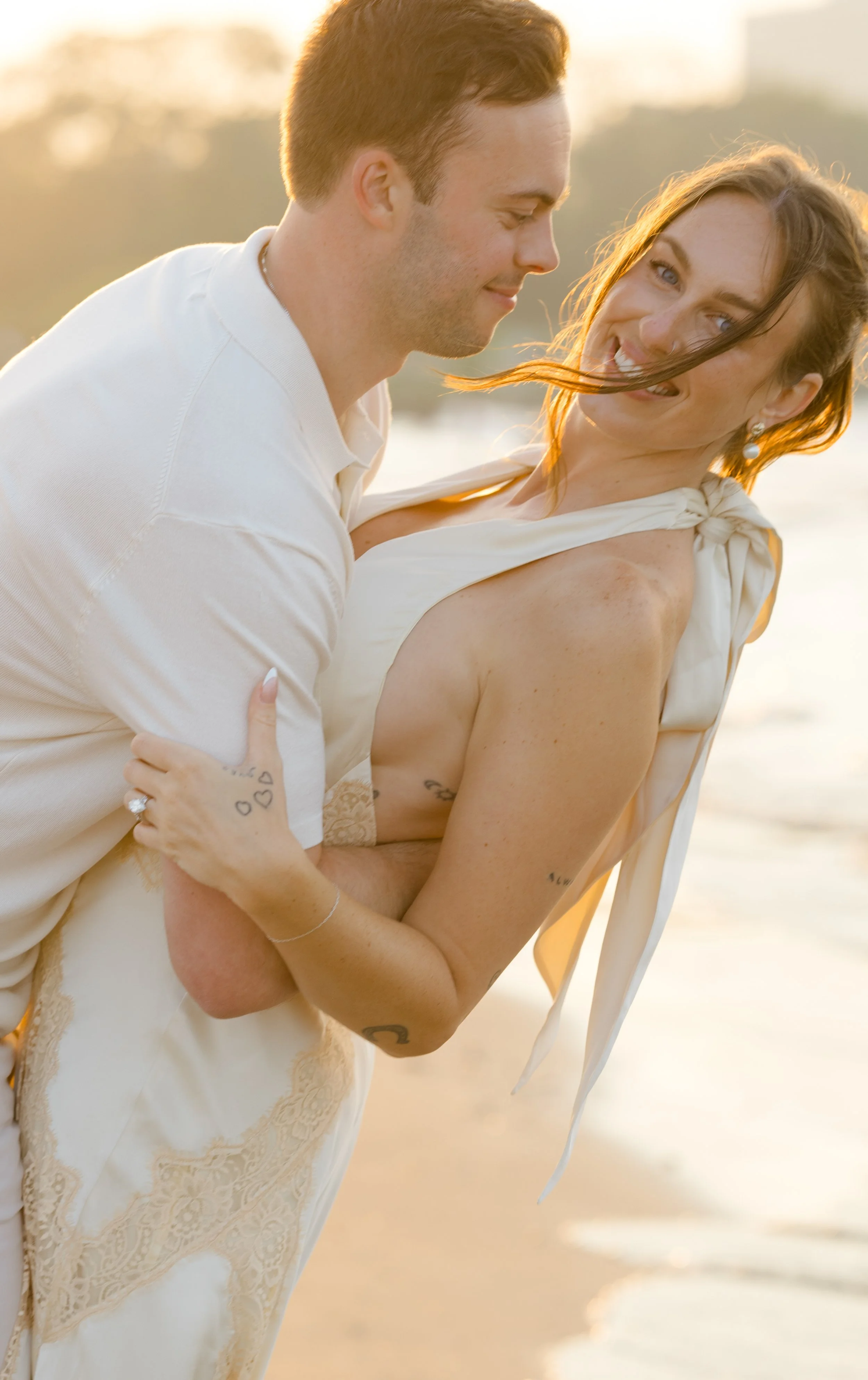 A man and woman smiling and embracing on a beach during sunset, with the ocean in the background.