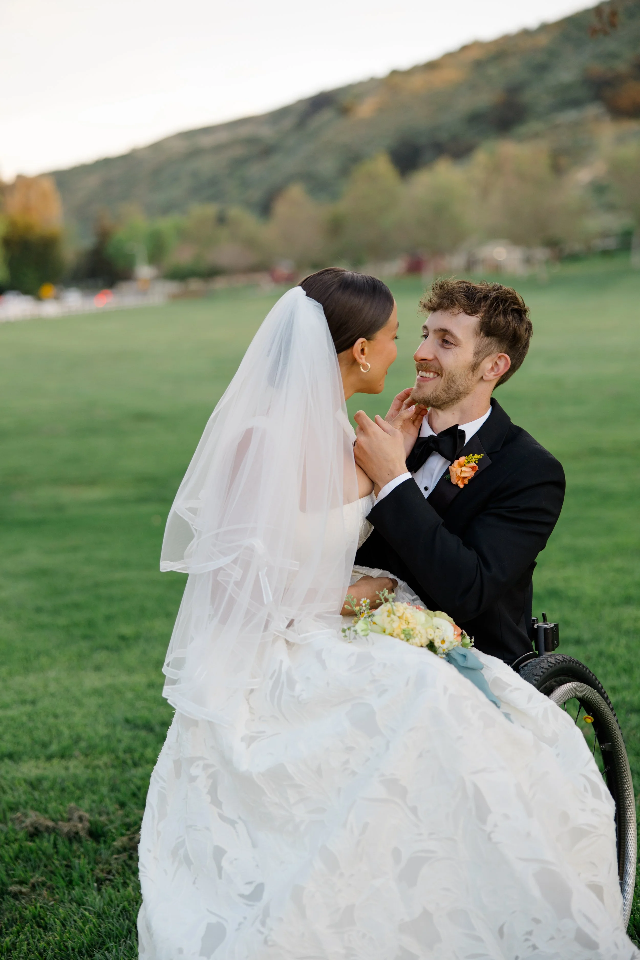 A bride with long dark hair in a white wedding gown and veil, and a groom in a black tuxedo, are smiling and gazing into each other's eyes outdoors in a grassy area with hills and trees in the background, during sunset.
