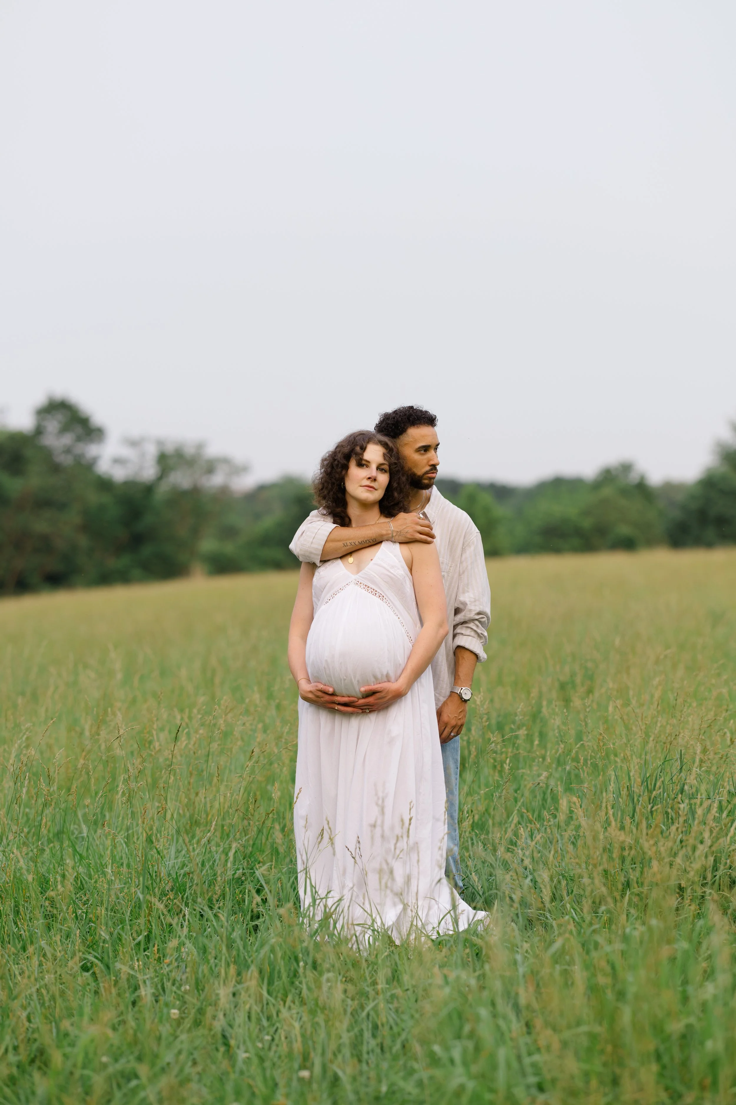 A pregnant woman with curly brown hair and a man with short curly hair standing in a grassy field with trees in the background. The woman is wearing a white dress and is cradling her belly, while the man stands behind her with his arm around her shou