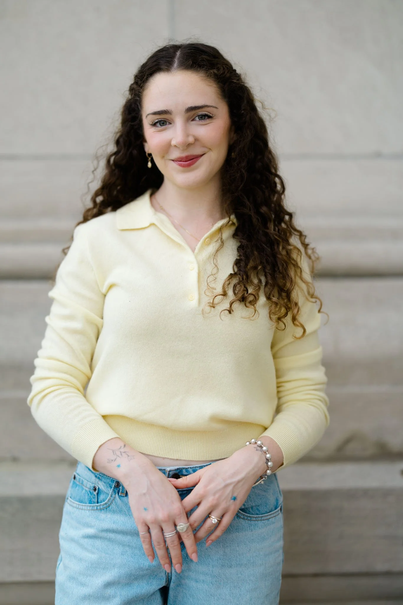 A young woman with long, curly brown hair and light skin standing in front of a gray concrete wall. She is wearing a pale yellow long-sleeve shirt and light blue jeans. She has multiple rings, a pearl bracelet, and small earrings. She is smiling soft