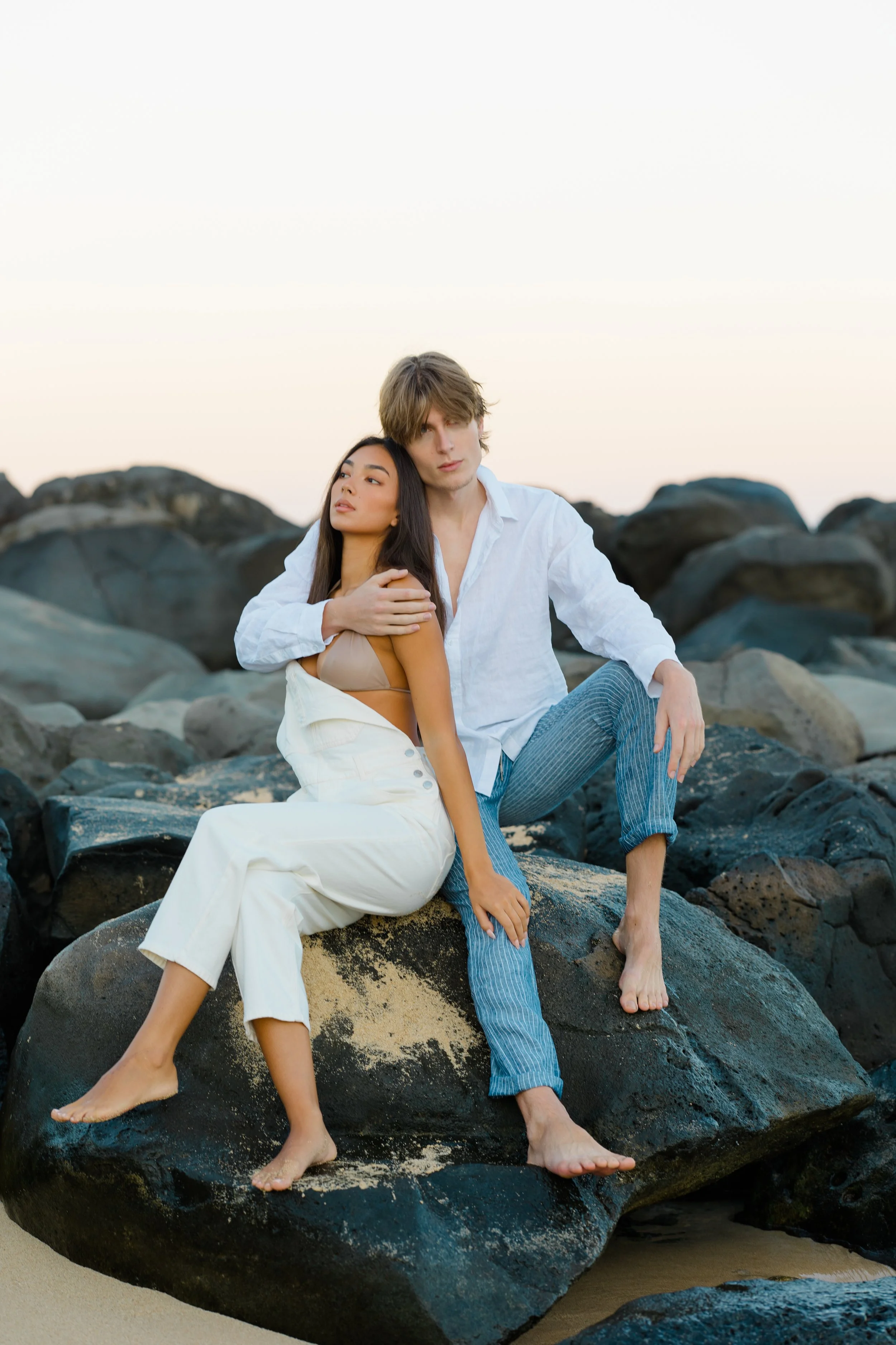 A man and woman sitting on large rocks at the beach during sunset, with the woman leaning on the man's shoulder. Both are barefoot, and the woman is wearing a beige top and white pants, while the man is dressed in a white shirt and blue striped pants.