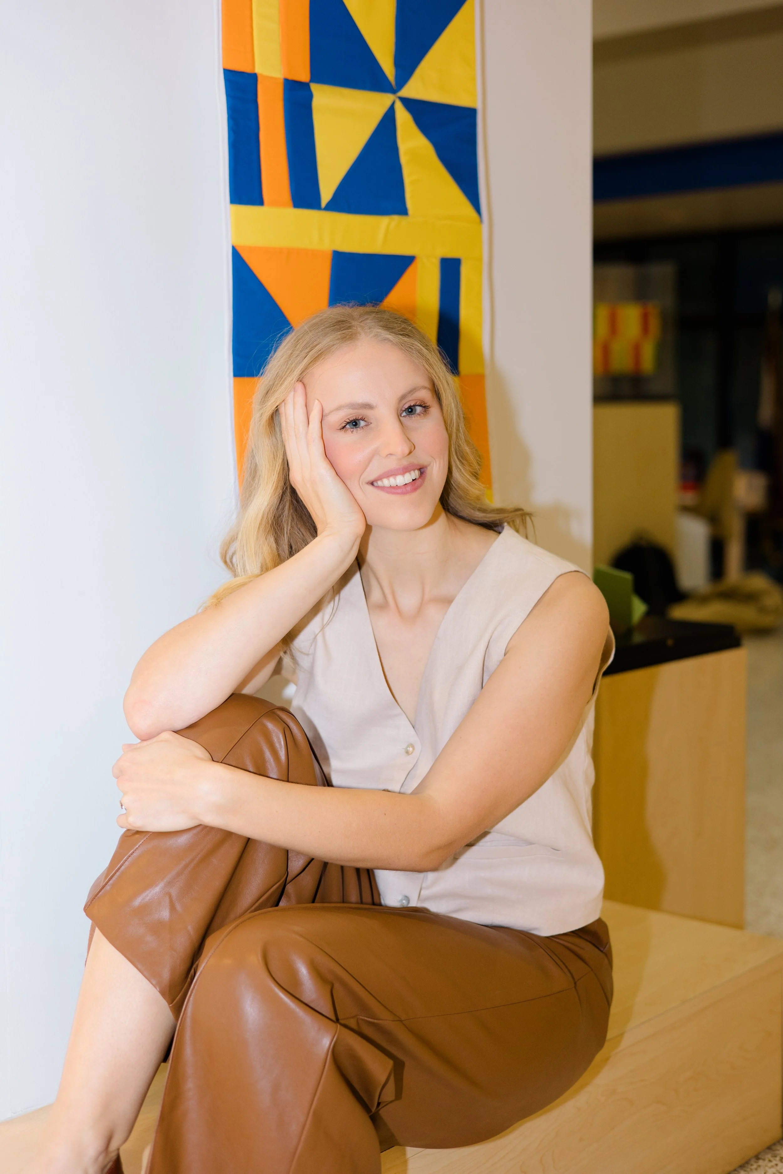 A young woman with blonde hair smiling, sitting on a brown leather armchair, resting her head on her left hand, with a colorful abstract quilt hanging on a white wall behind her.