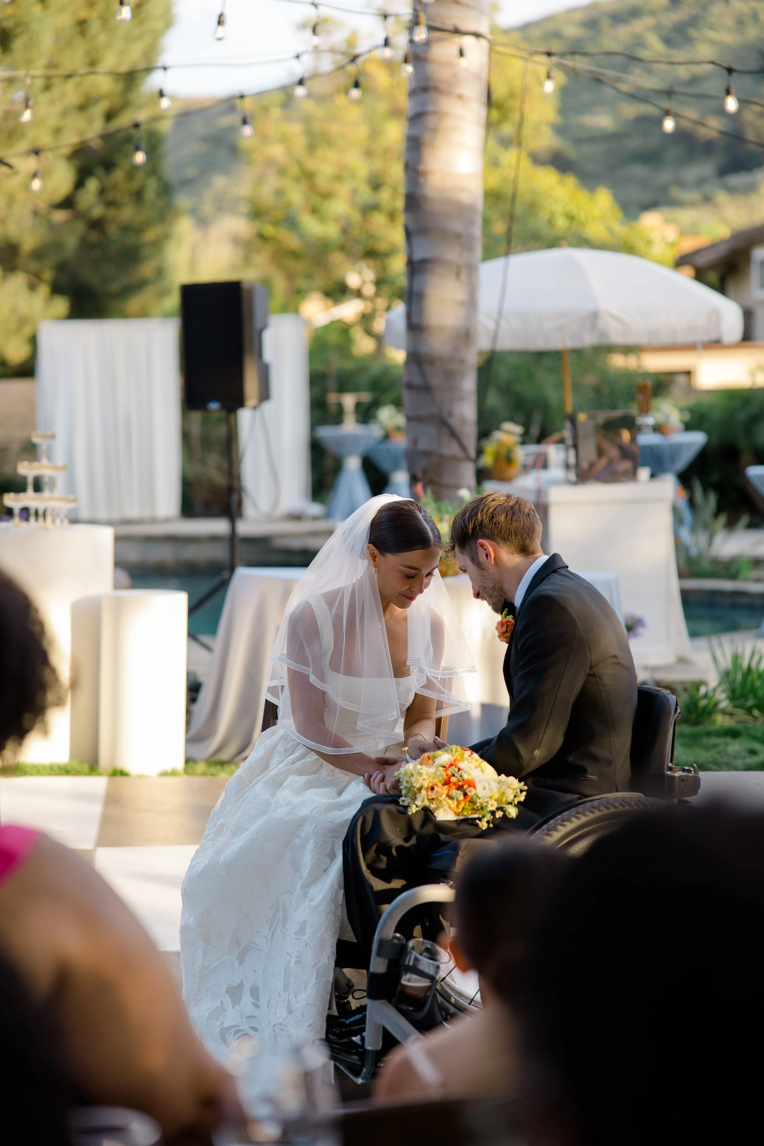 A bride and groom kneeling together during their wedding ceremony outdoors, with trees and string lights in the background, a large white umbrella, and a decorated table.