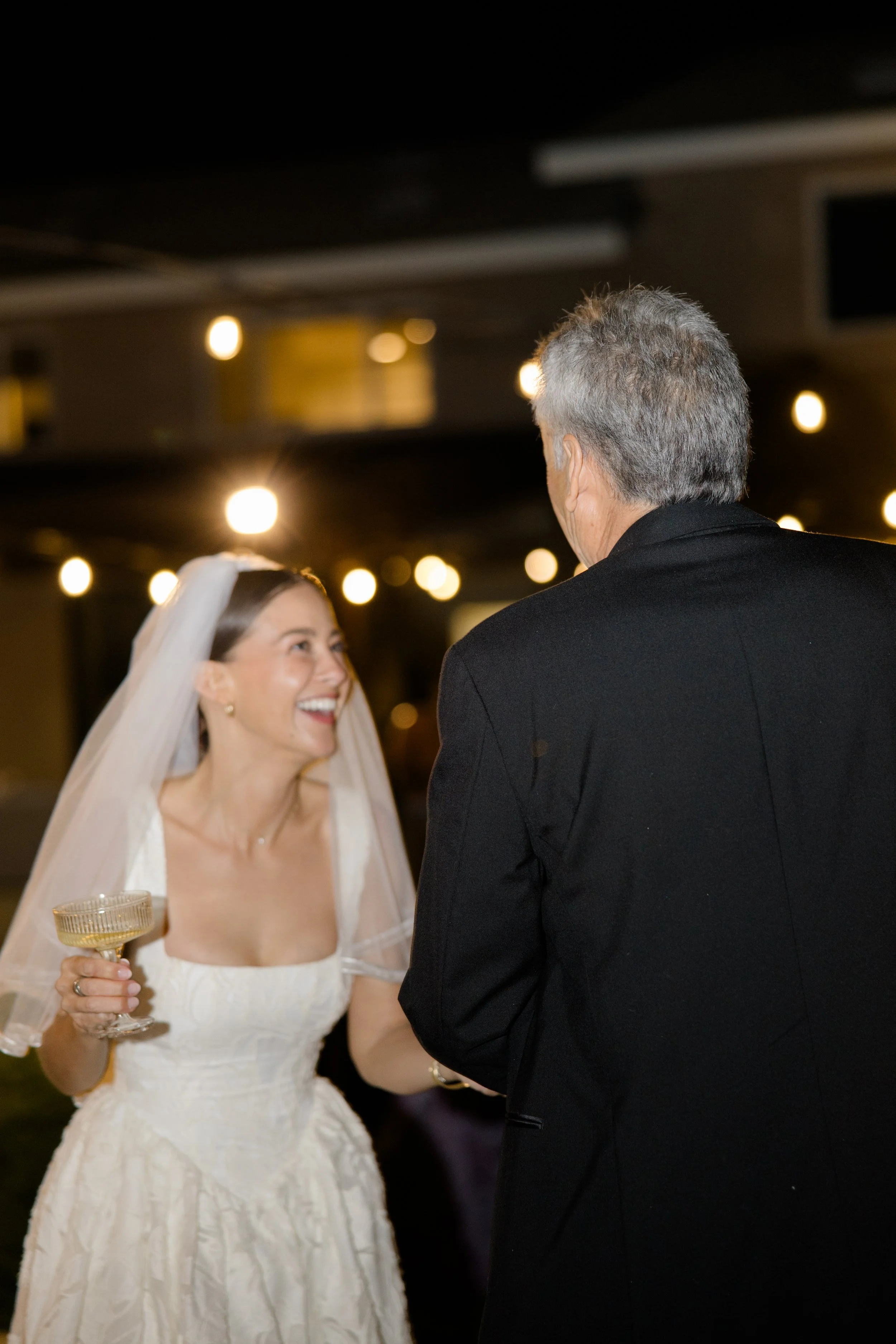 A bride with a white wedding dress and veil holding a champagne glass, smiling and looking at an older man in a black suit, in a dimly lit reception hall with string lights.
