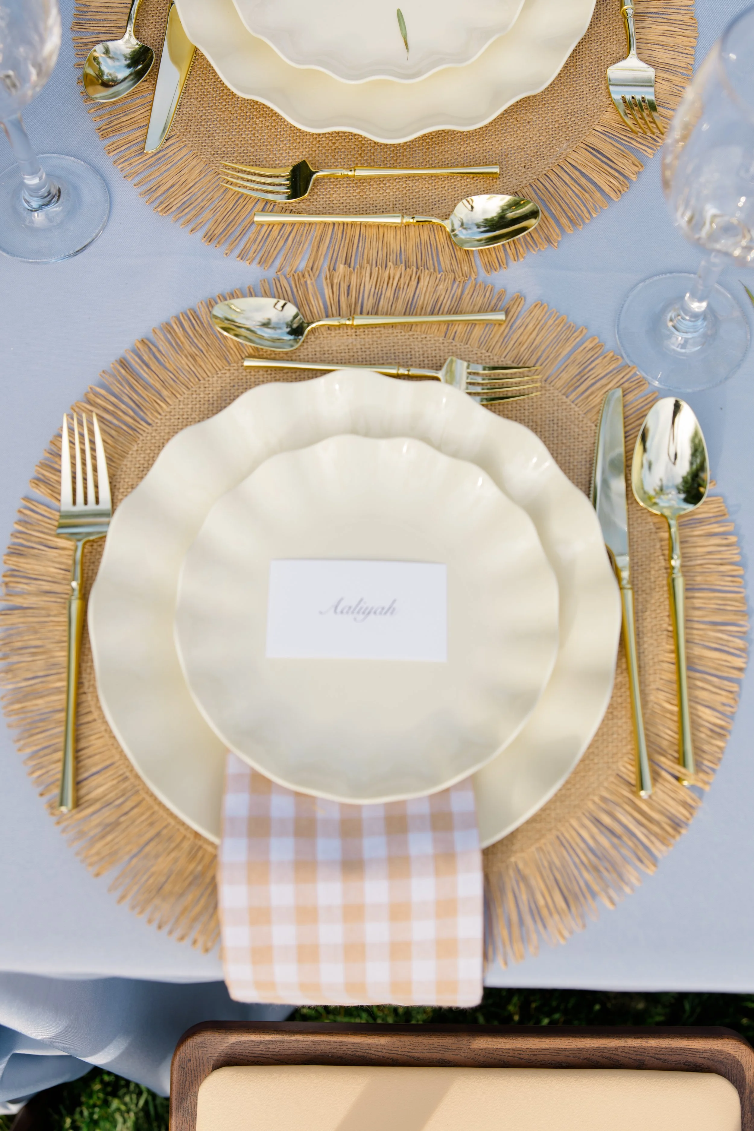 Table setting with white plates, gold utensils, clear wine glasses, a white salad bowl, a burlap placemat, and a blue tablecloth. There is a name card on the table.