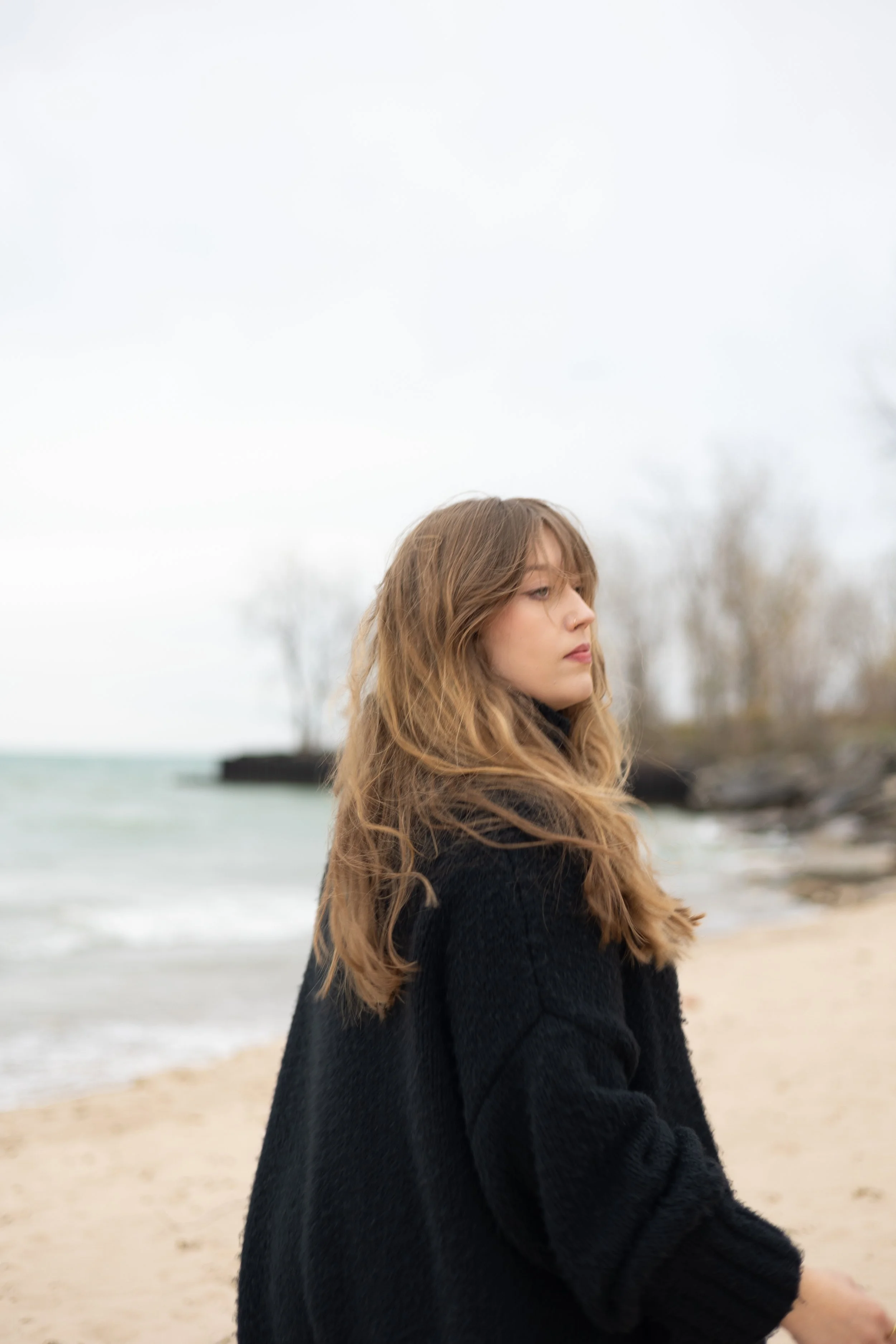 A young woman with long wavy hair wearing a black coat, standing on a beach with water, sand, and trees in the background on a cloudy day.