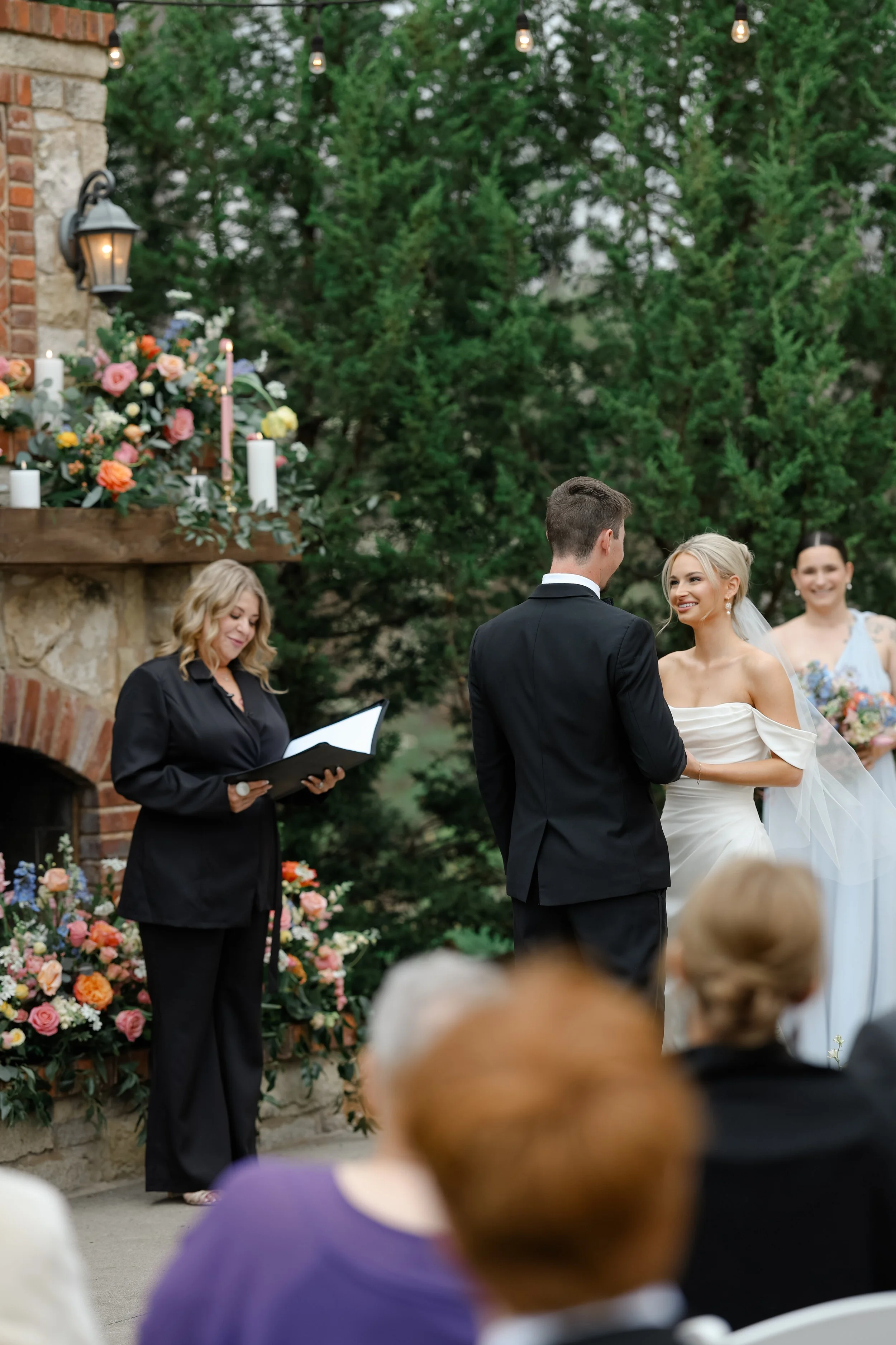 An outdoor wedding ceremony with a bride and groom facing each other, surrounded by guests. The ceremony takes place in front of a floral arrangement with pink, orange, and white flowers, candles, and greenery. There are string lights hanging overhea