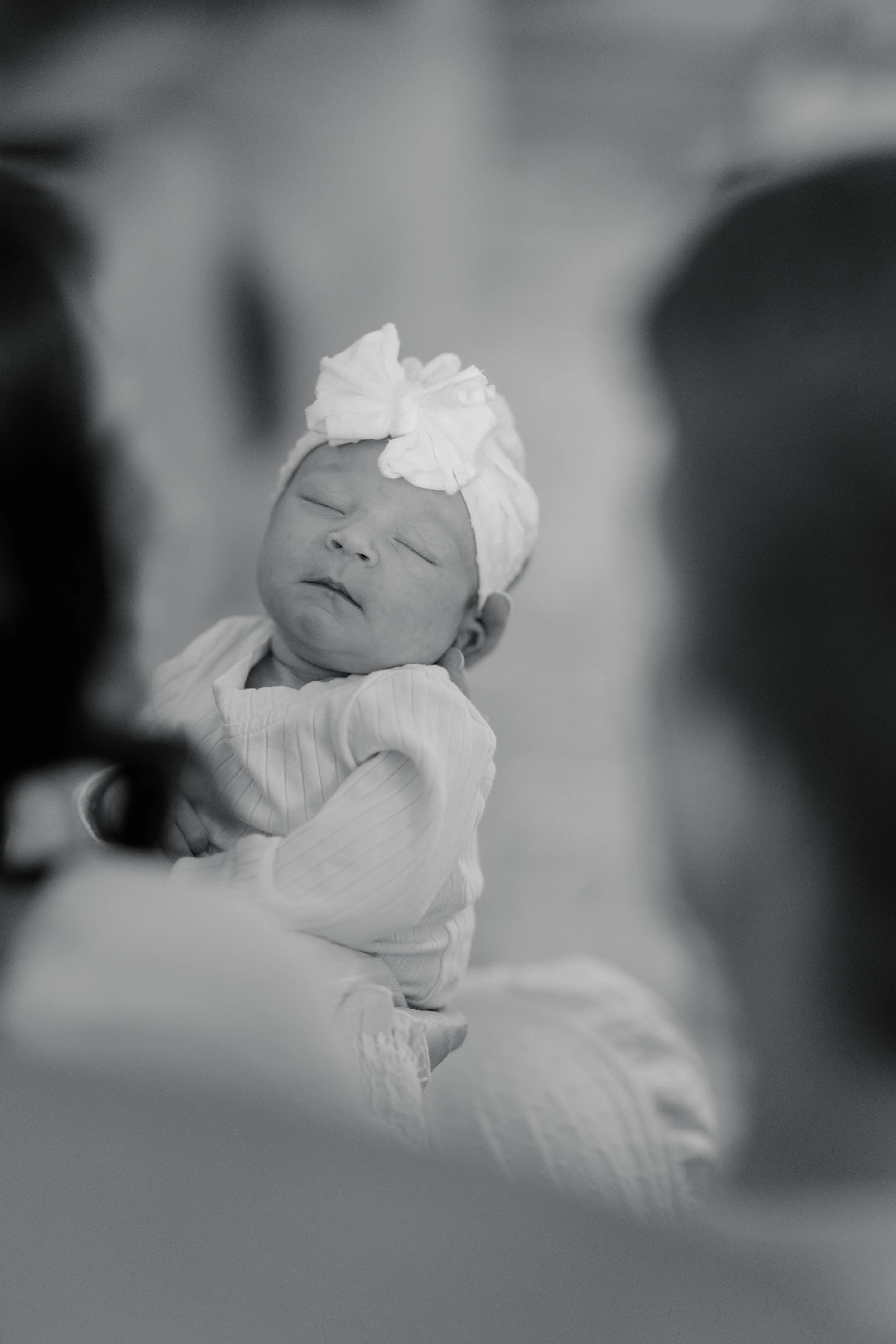 A black and white photo of a newborn baby wearing a headband with a bow, being held by an adult, with blurred people in the background.