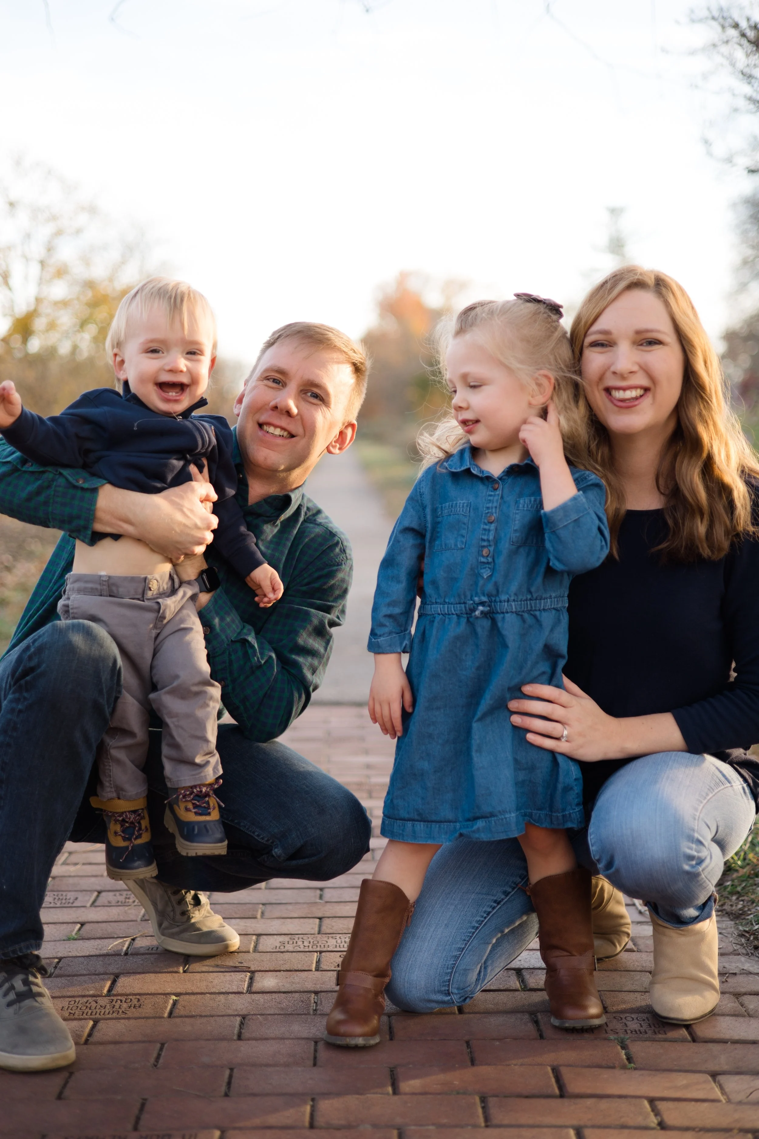 A family of four with two young children, a boy and a girl, outdoors during daytime. The father is kneeling and holding the boy, who is smiling and wearing a navy jacket. The mother is kneeling beside the girl, who is wearing a denim dress and boots.