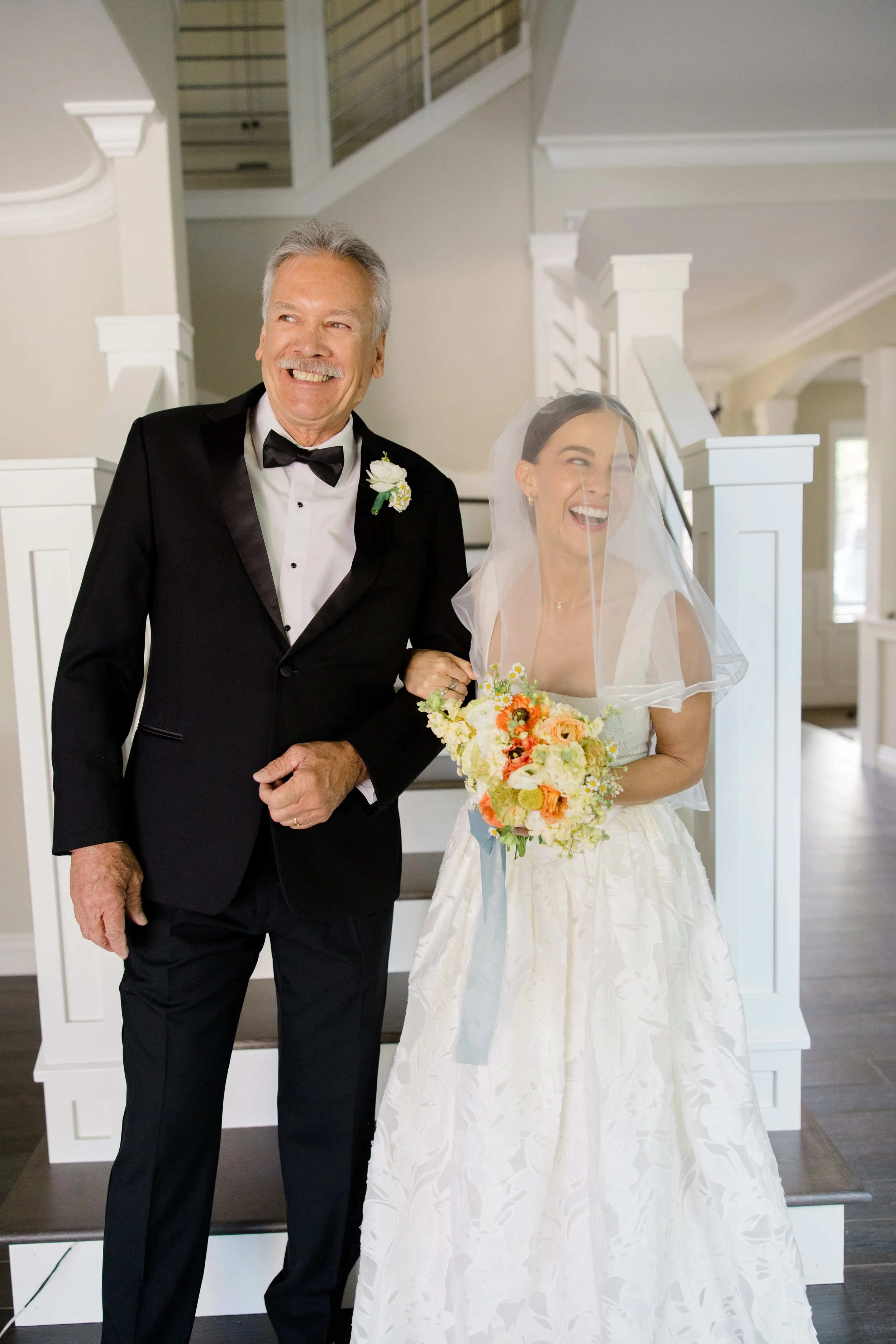 An elderly man in a tuxedo and black bow tie smiling next to a bride in a white wedding dress and veil holding a colorful bouquet of flowers. They are standing indoors near a staircase.