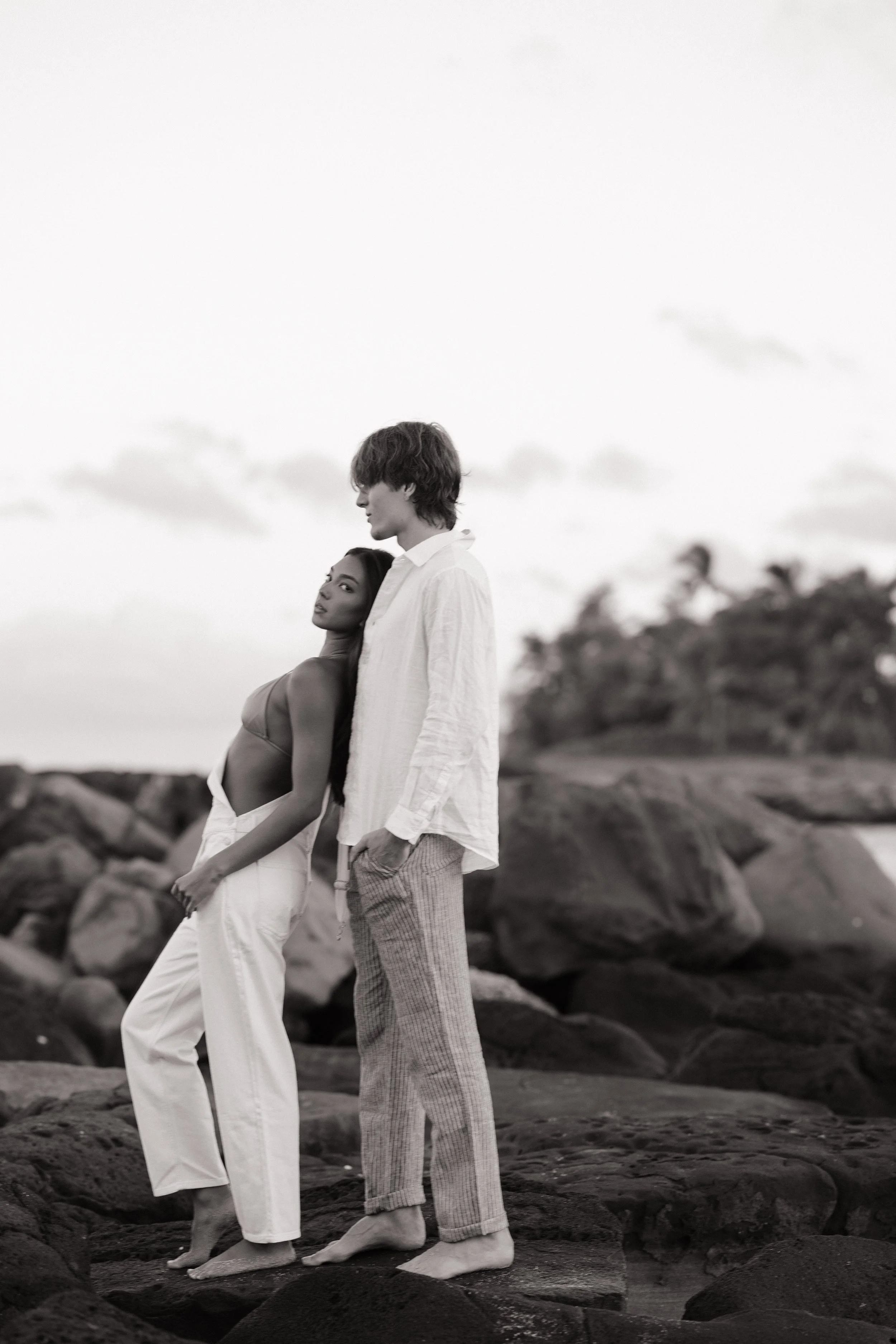 A black and white photo of a young man and woman standing on rocks near a beach or shoreline during daytime. The woman is leaning slightly against the man, and they are both looking in different directions. The woman is wearing a sleeveless top and w