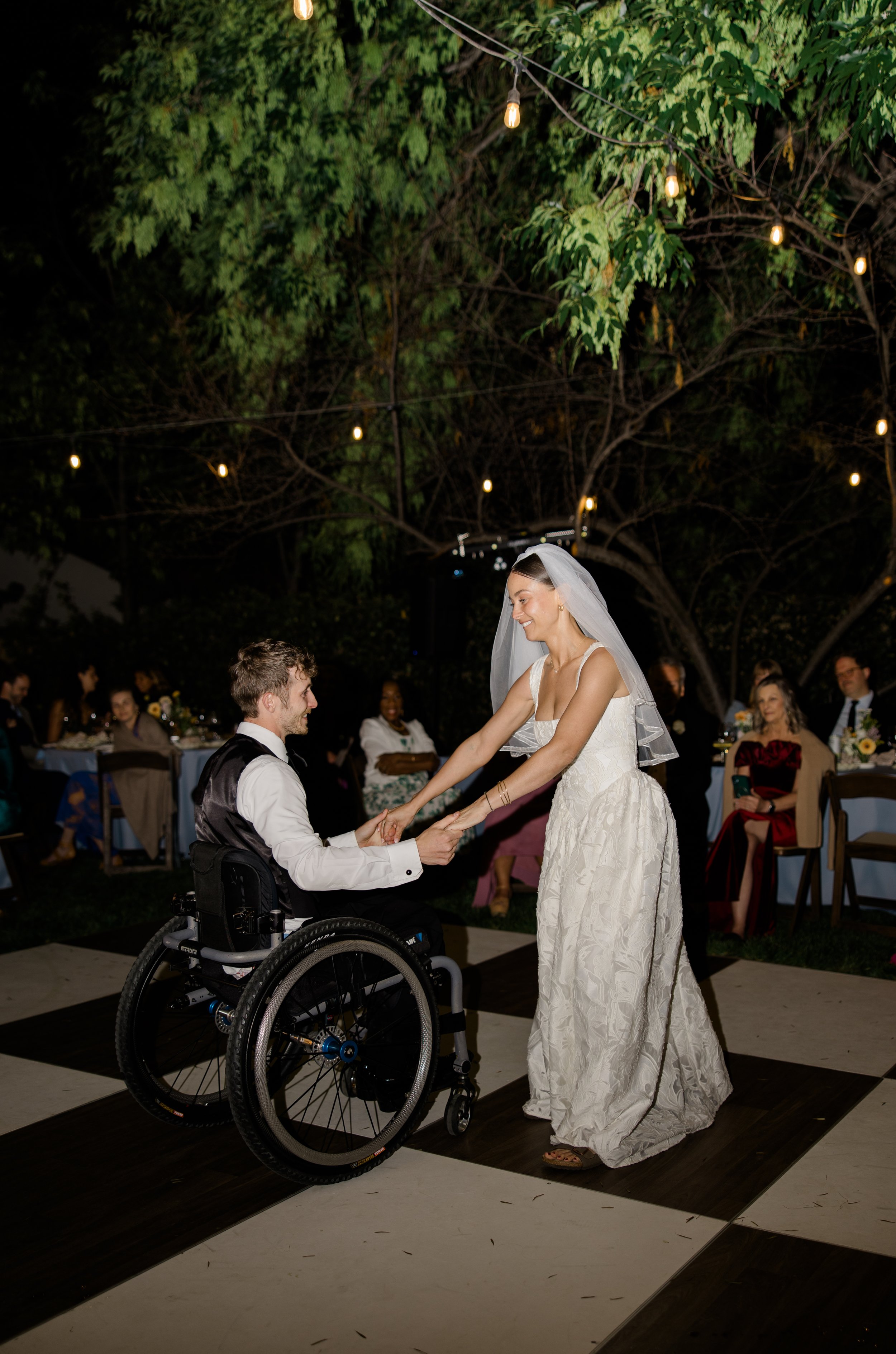 A bride and groom at a wedding reception, with the groom in a wheelchair. They are holding hands and smiling at each other, surrounded by guests seated at tables in an outdoor setting decorated with string lights and a large tree.
