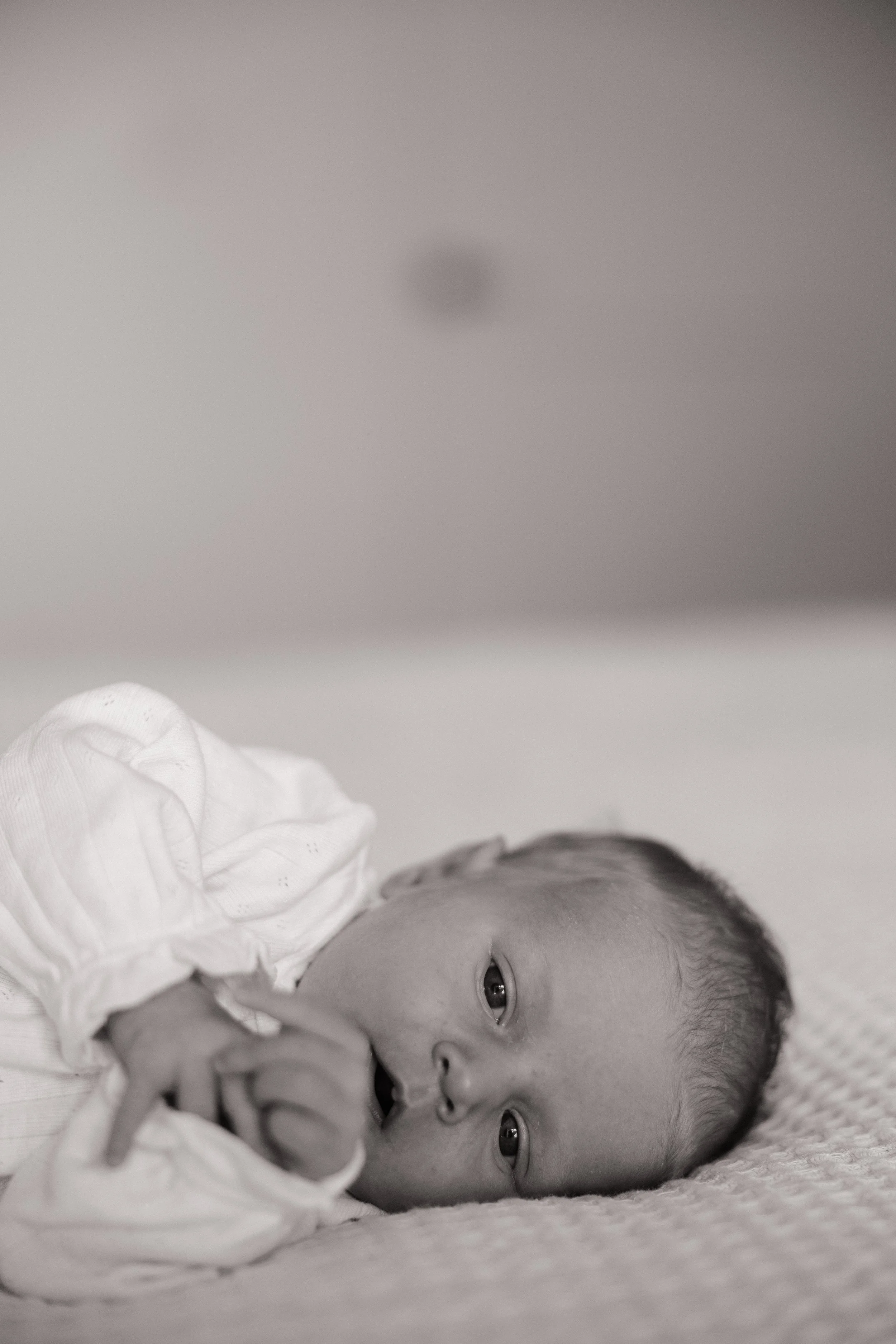 A baby lying on a soft surface, dressed in a white outfit, with a focused expression, looking towards the camera, with a plain wall background.