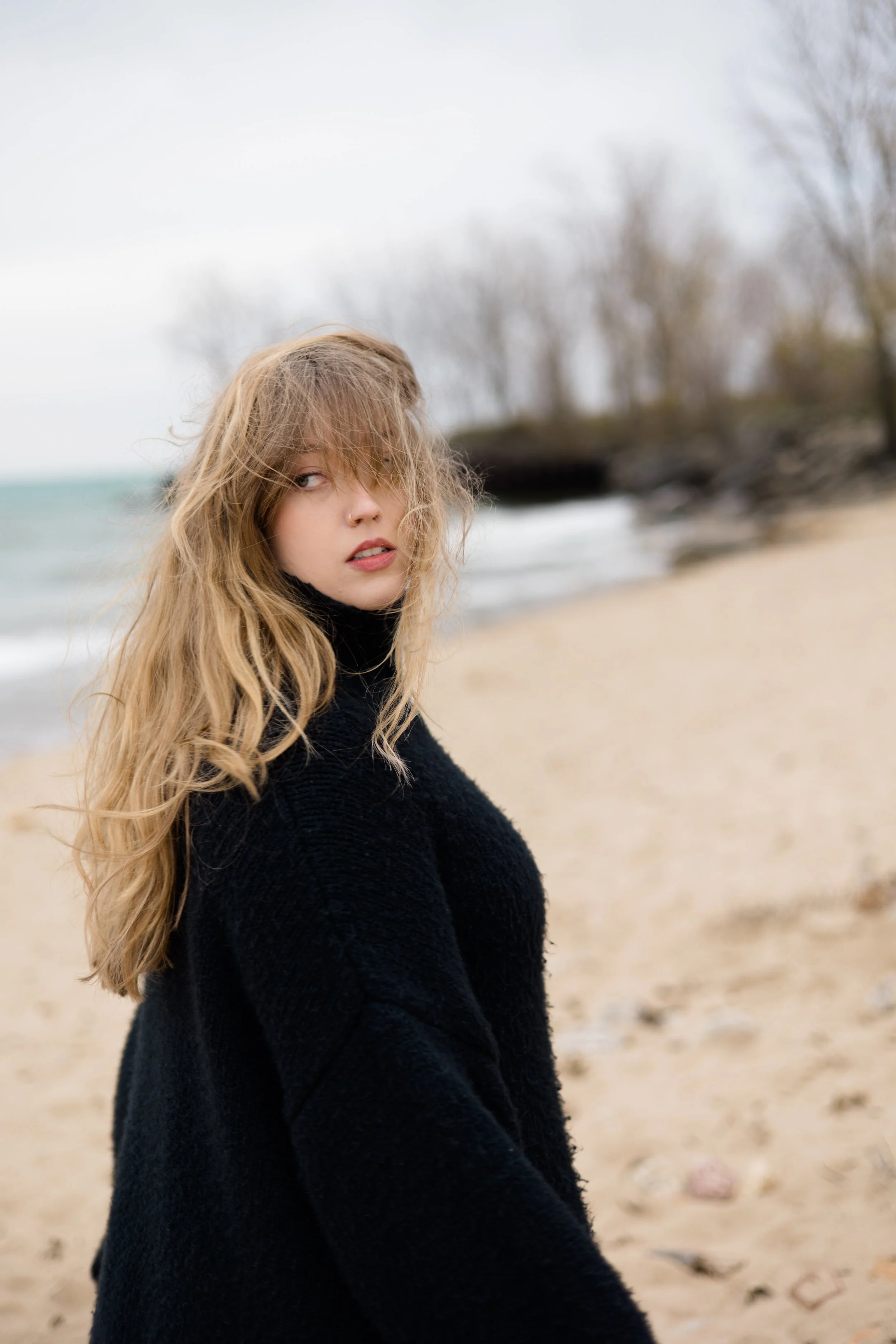 A young woman with long, wavy blonde hair and fair skin, wearing a black sweater, standing on a beach with an overcast sky and a blurred background of trees and rocks.