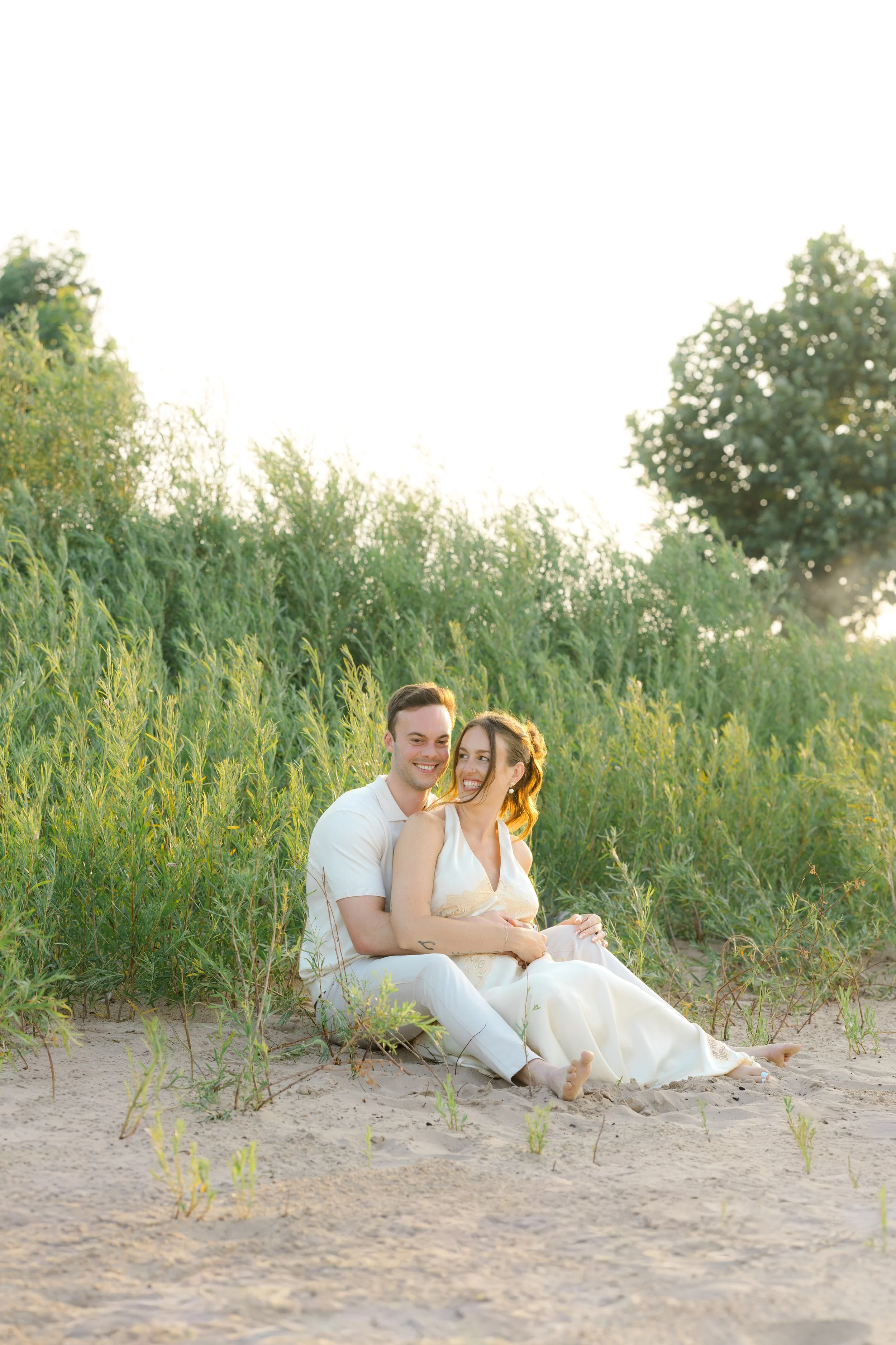 A happy couple sitting on the sand, embracing each other, surrounded by tall green grass and trees at sunset.