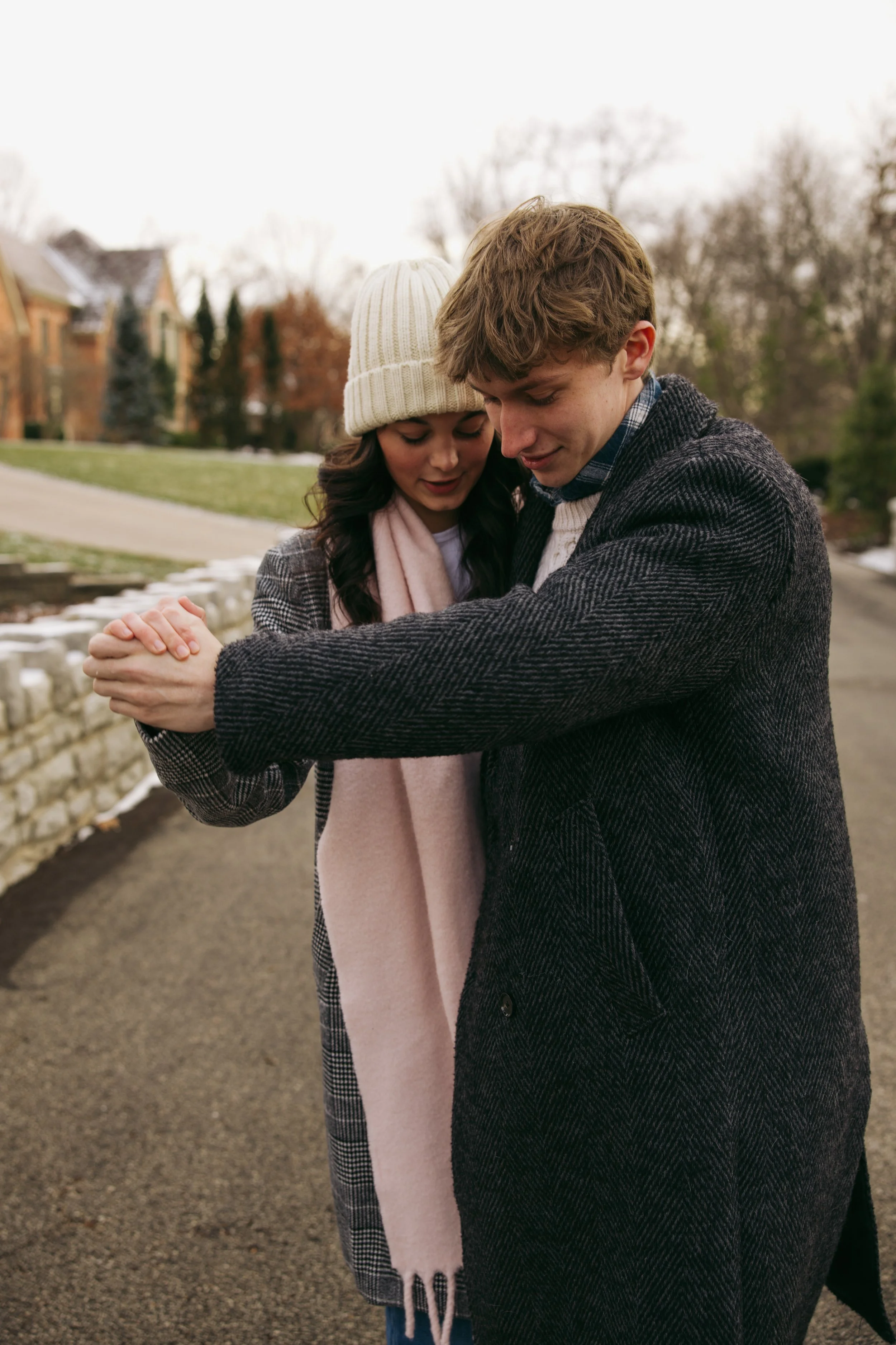 A young couple is standing together outdoors on a cloudy day, embracing with their heads bowed towards each other and eyes closed. The woman is wearing a cream knit beanie and a gray coat, while the man has curly brown hair and is dressed in a dark g