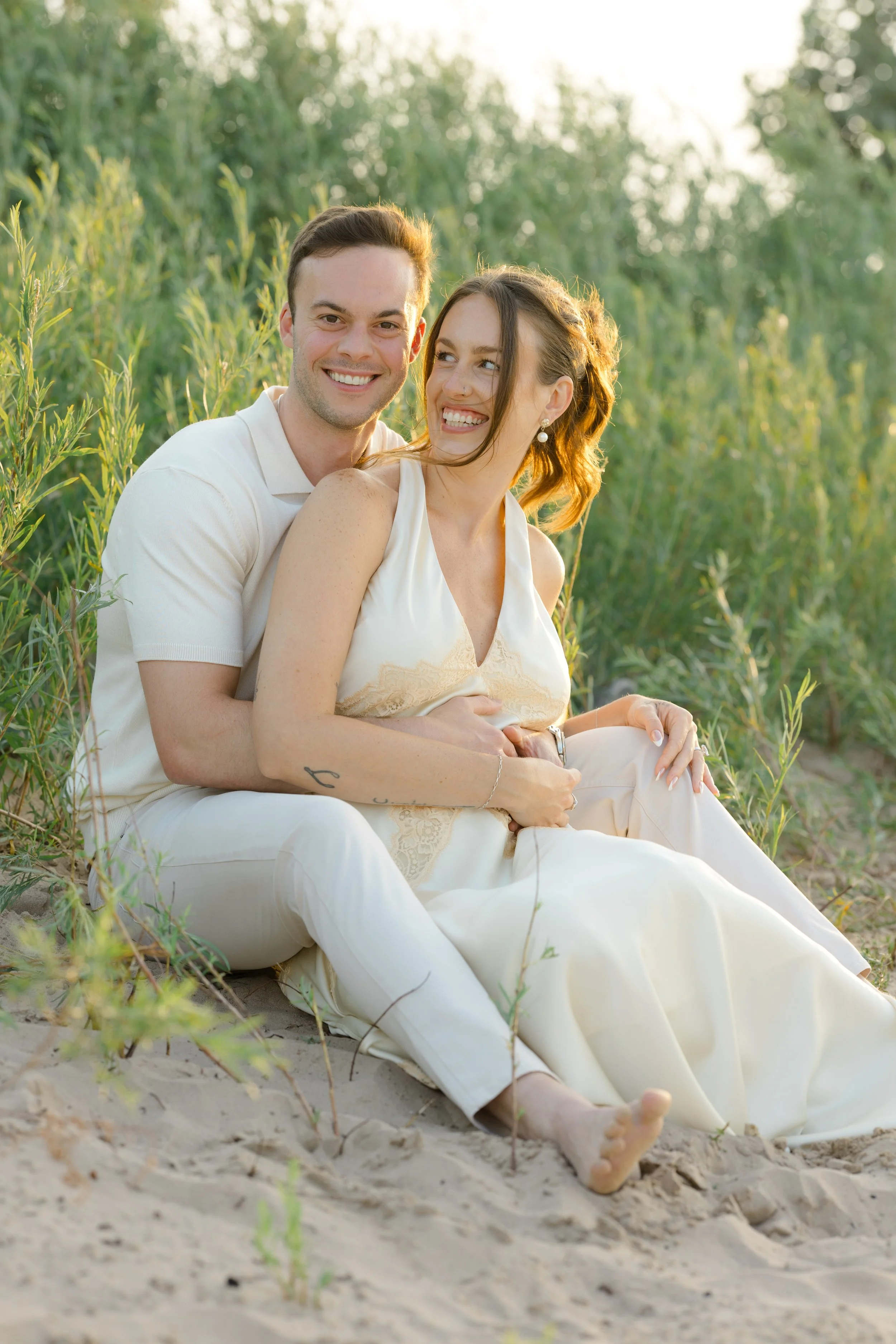 A smiling man and woman sitting on the sandy ground in a field with green plants, during sunset or late afternoon, close together, the woman looking at the man.