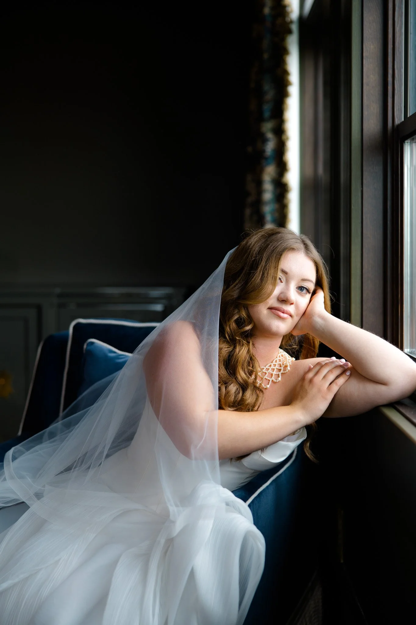 A young woman with long wavy red hair, wearing a white wedding dress and veil, sitting by a window with natural light, looking at the camera with a gentle expression, inside a cozy room with dark walls and a window with dark wooden frames.