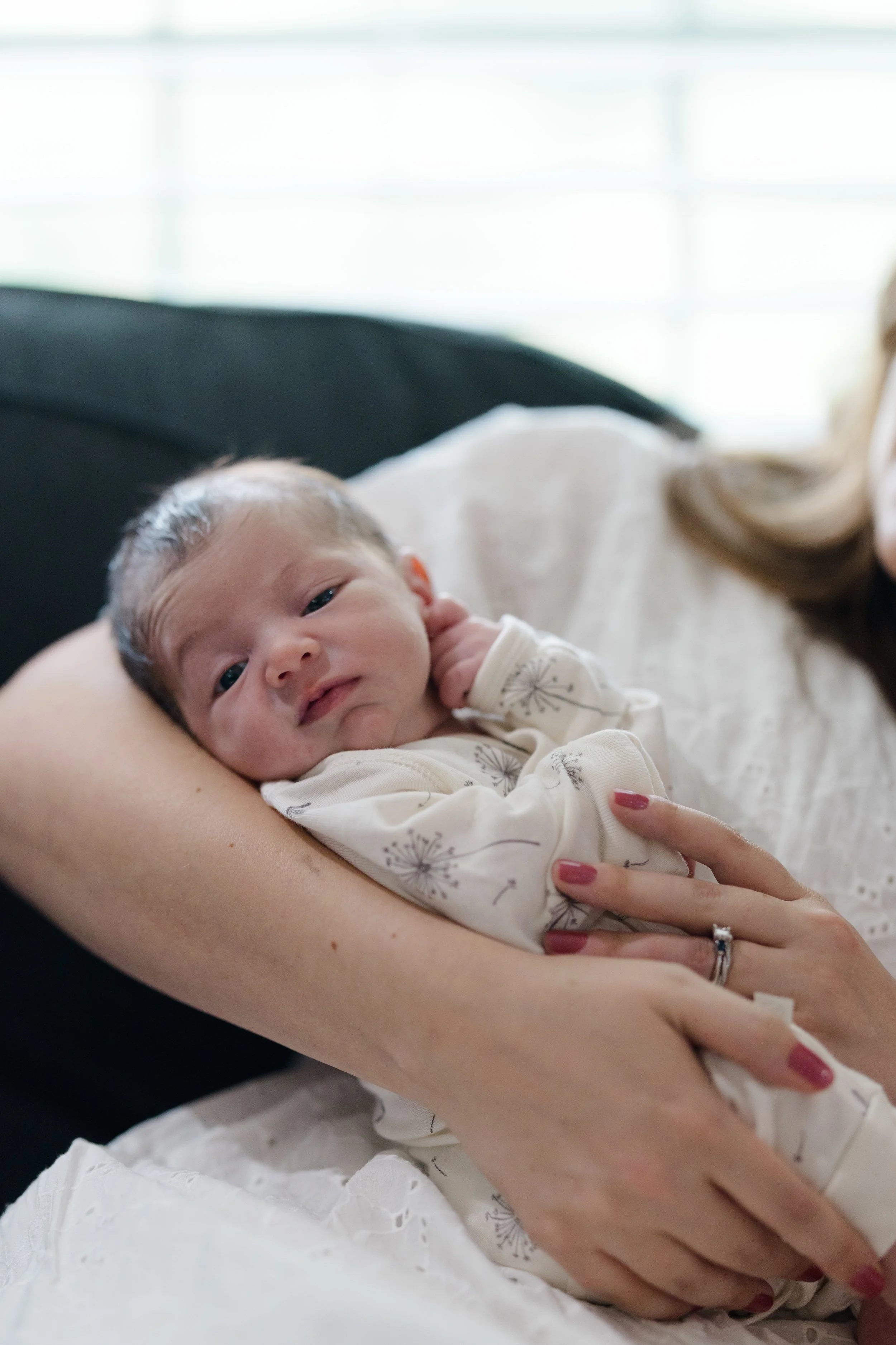A newborn baby lying in a person's arms, wearing a white outfit with dandelion print, with an adult’s hand supporting the baby, visible nails painted pink, and a ring on the ring finger. The background is softly lit with a blurred window.