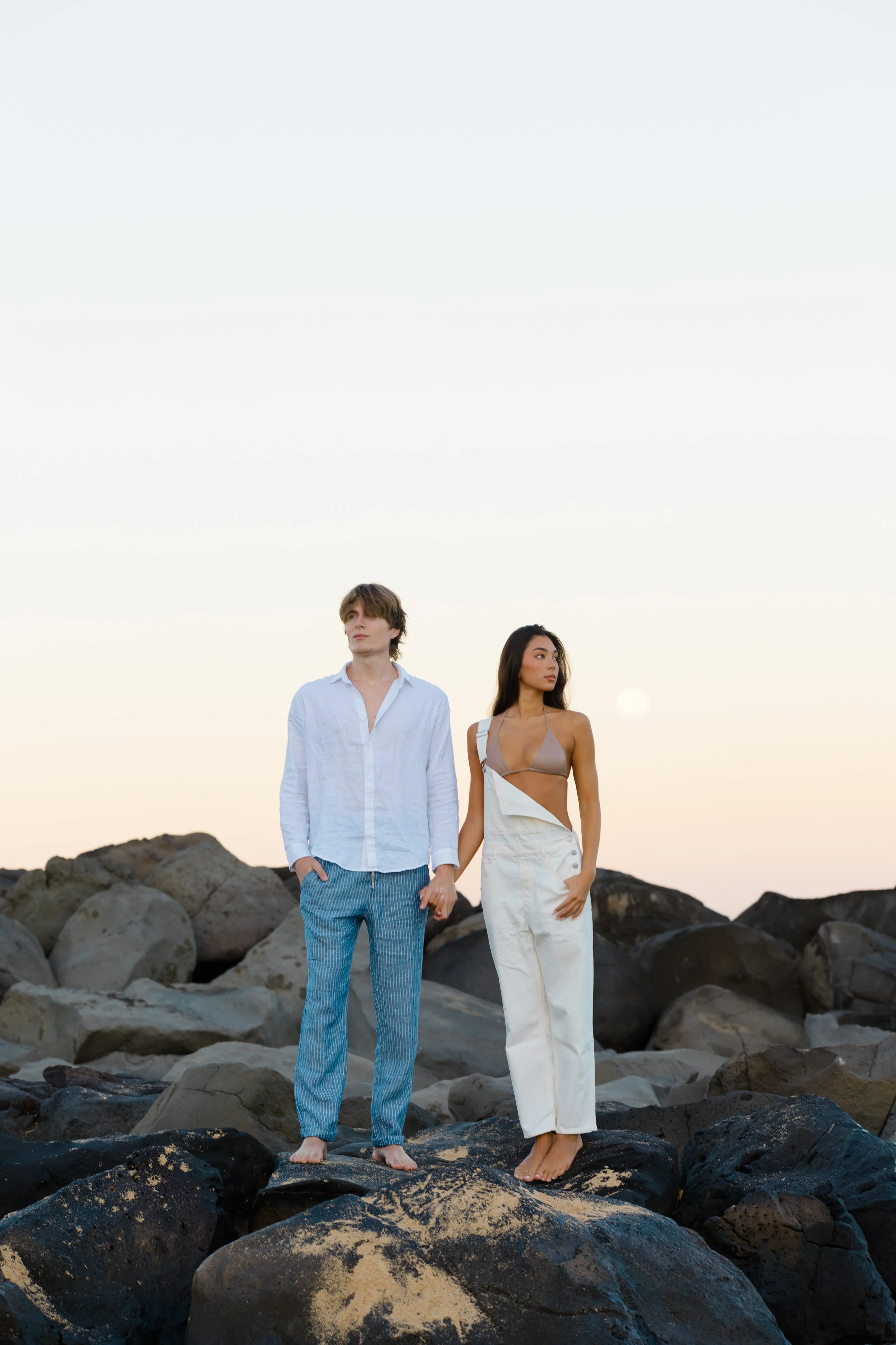 A man and woman standing barefoot on large rocks at the beach during sunset, holding hands, with the sky in the background.