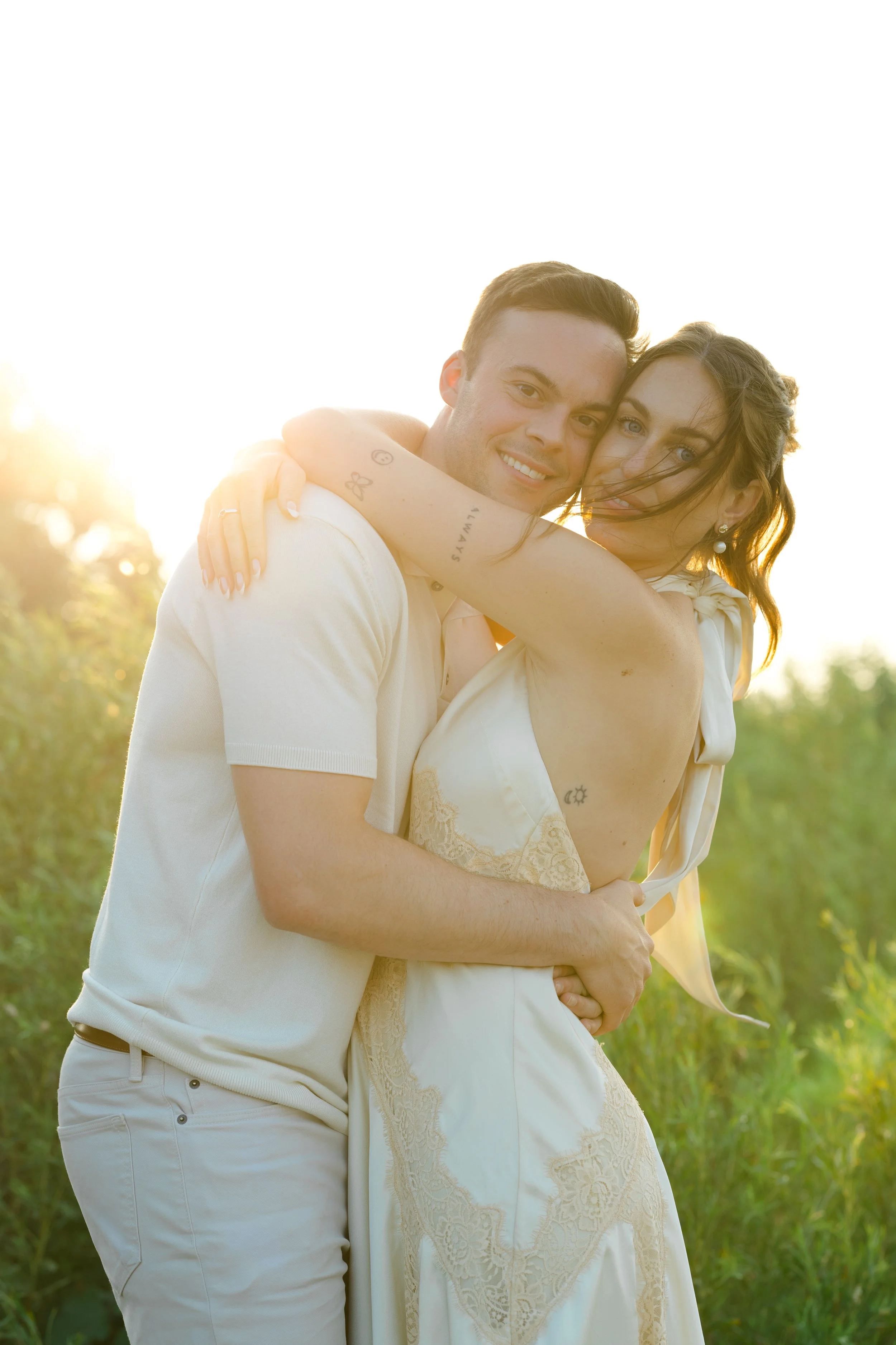 A smiling couple, a man and a woman, embracing outdoors with sunlight behind them. The man is wearing a white polo shirt, and the woman is wearing a light-colored dress with lace details. The woman has tattoos on her arm and shoulder, and has wind-bl