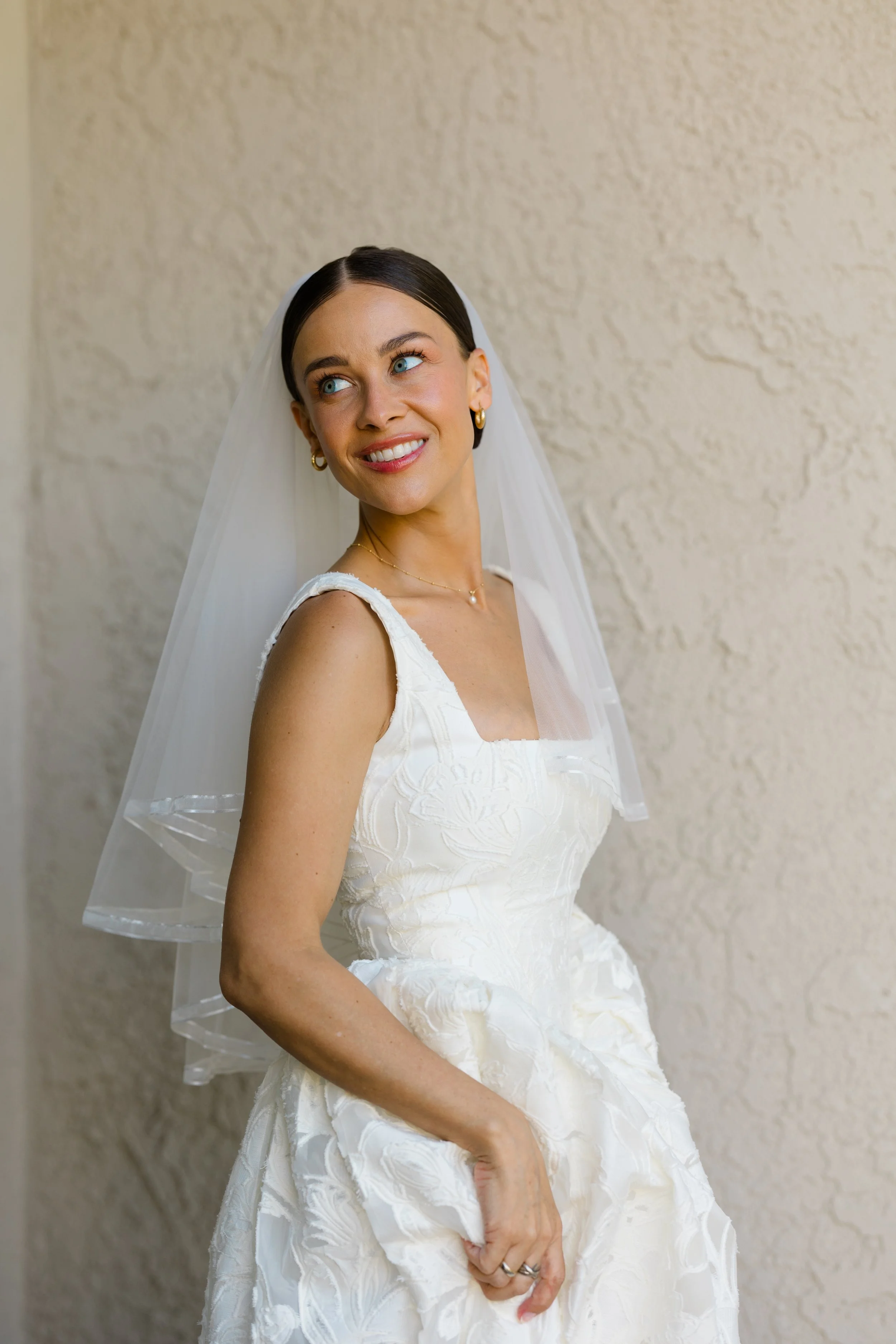 A woman wearing a white wedding dress and veil is smiling and looking to her left. She is standing against a textured light-colored wall.