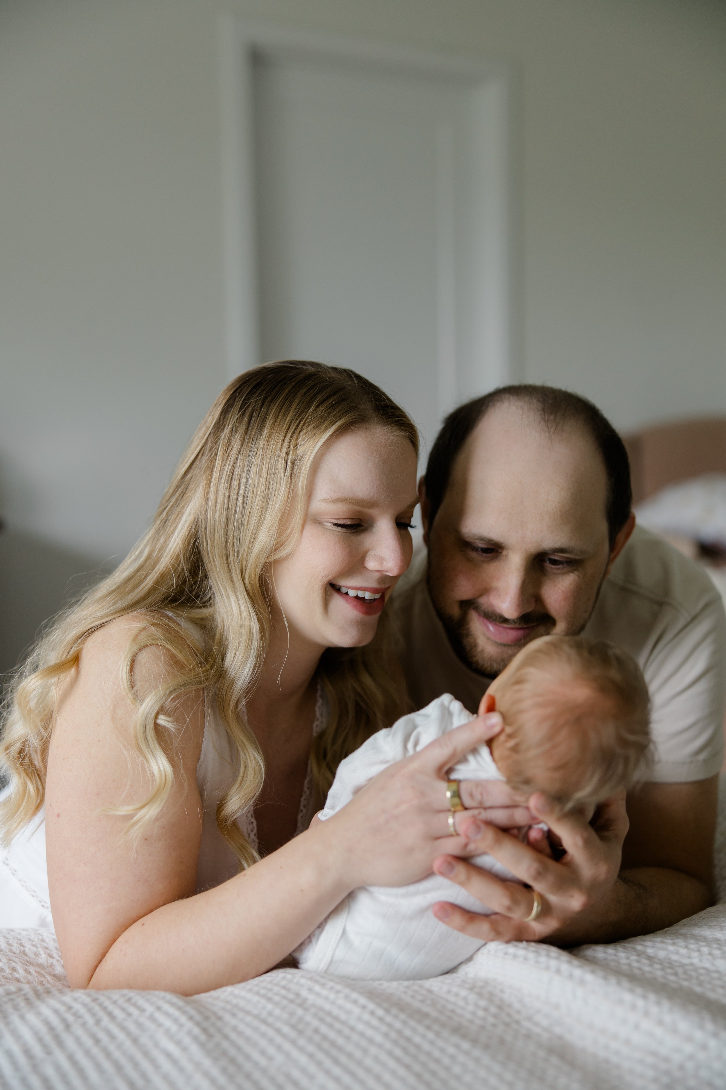A young woman with long blonde hair, smiling, holding a baby, with a man next to her also smiling and looking at the baby, in a cozy room.