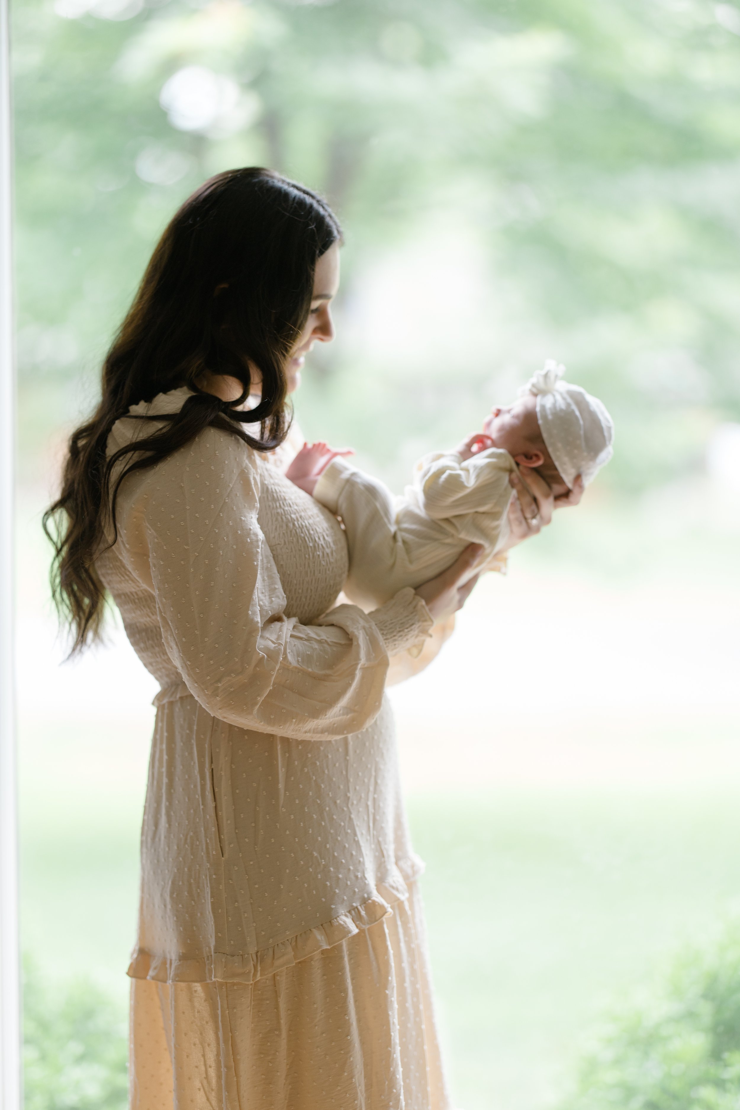 A woman with long dark hair, dressed in a beige dress with lace details, is holding a baby dressed in cream, with a matching beanie. They are indoors near a window with a blurred green outdoor background.